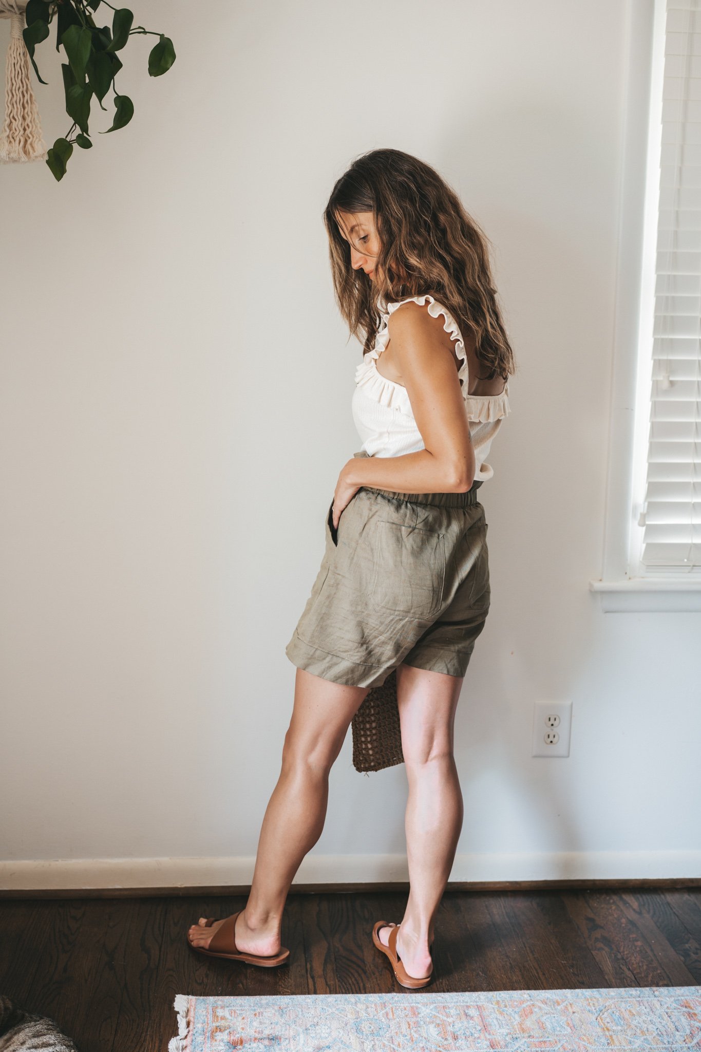 ashley wearing a cream colored tank with ruffled sleeves, olive green paperbag shorts, brown leather sandals, and a woven bag