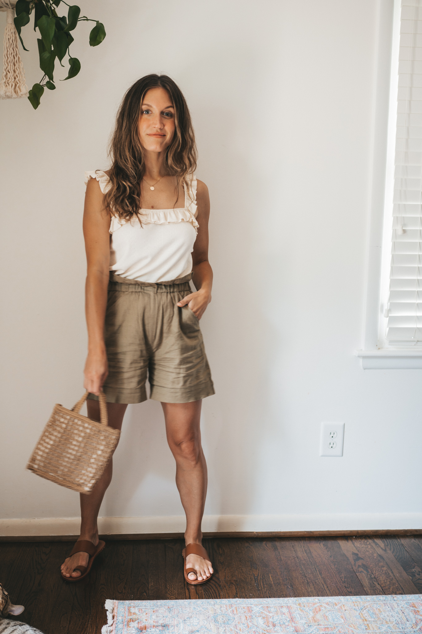 ashley wearing a cream colored tank with ruffled sleeves, olive green paperbag shorts, brown leather sandals, and a woven bag