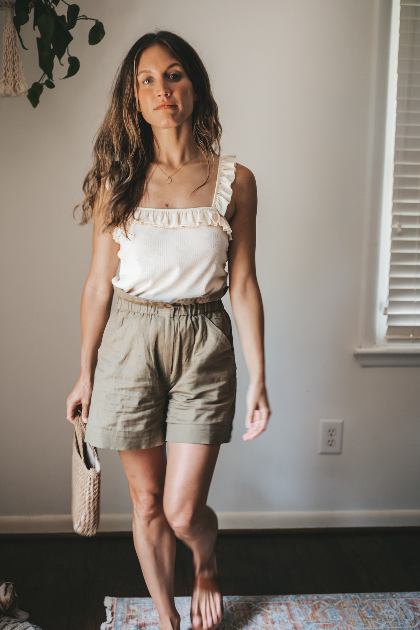 ashley wearing a cream colored tank with ruffled sleeves, olive green paperbag shorts, brown leather sandals, and a woven bag