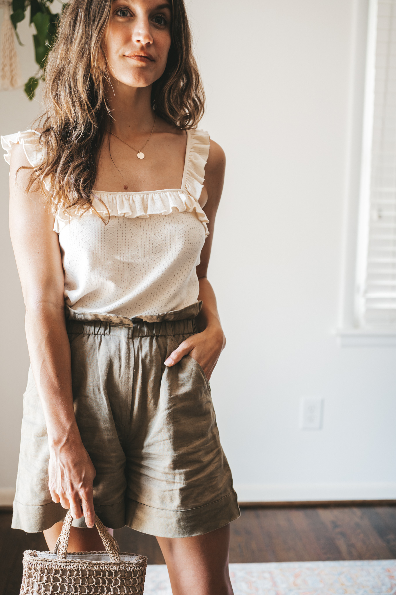ashley wearing a cream colored tank with ruffled sleeves, olive green paperbag shorts, brown leather sandals, and a woven bag