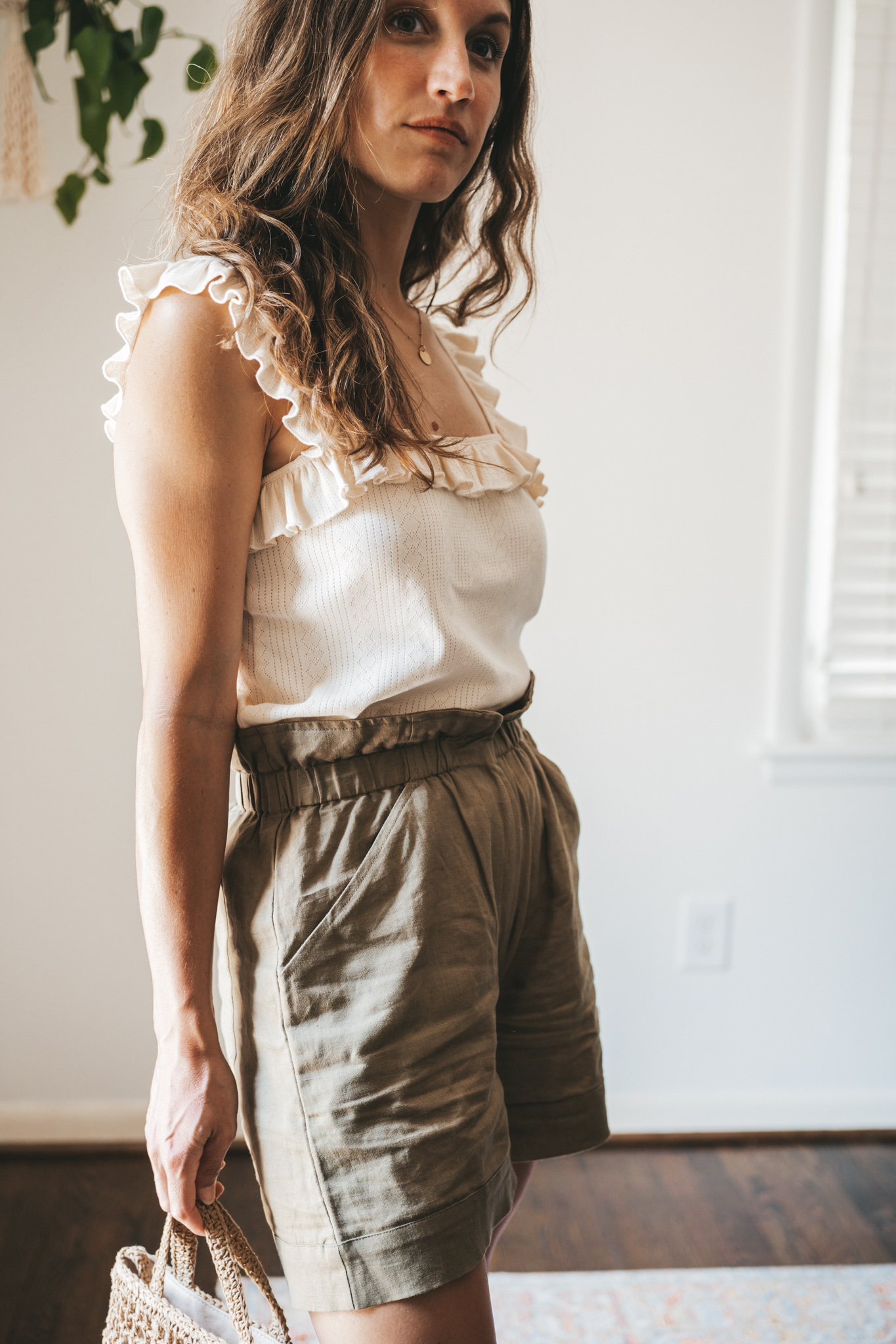 ashley wearing a cream colored tank with ruffled sleeves, olive green paperbag shorts, brown leather sandals, and a woven bag