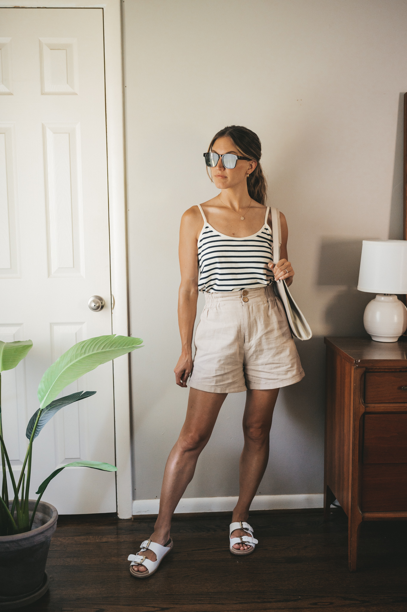 ashley wearing a white and navy striped tank, tan linen shorts, white sandals, a gray shoulder bag, and tortoise shell sunglasses