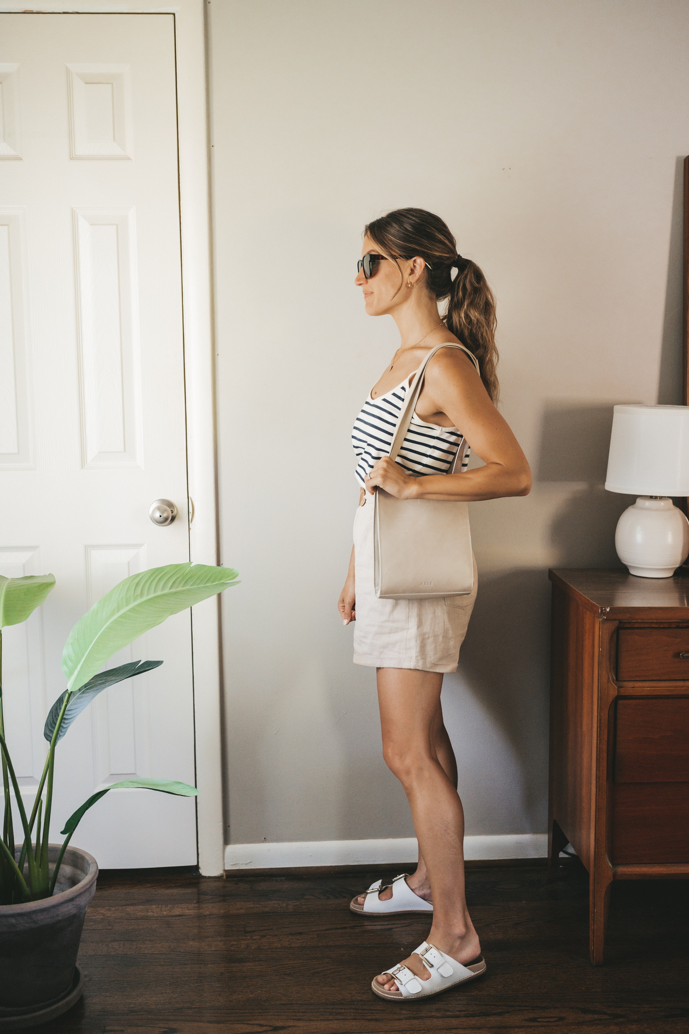 ashley wearing a white and navy striped tank, tan linen shorts, white sandals, a gray shoulder bag, and tortoise shell sunglasses