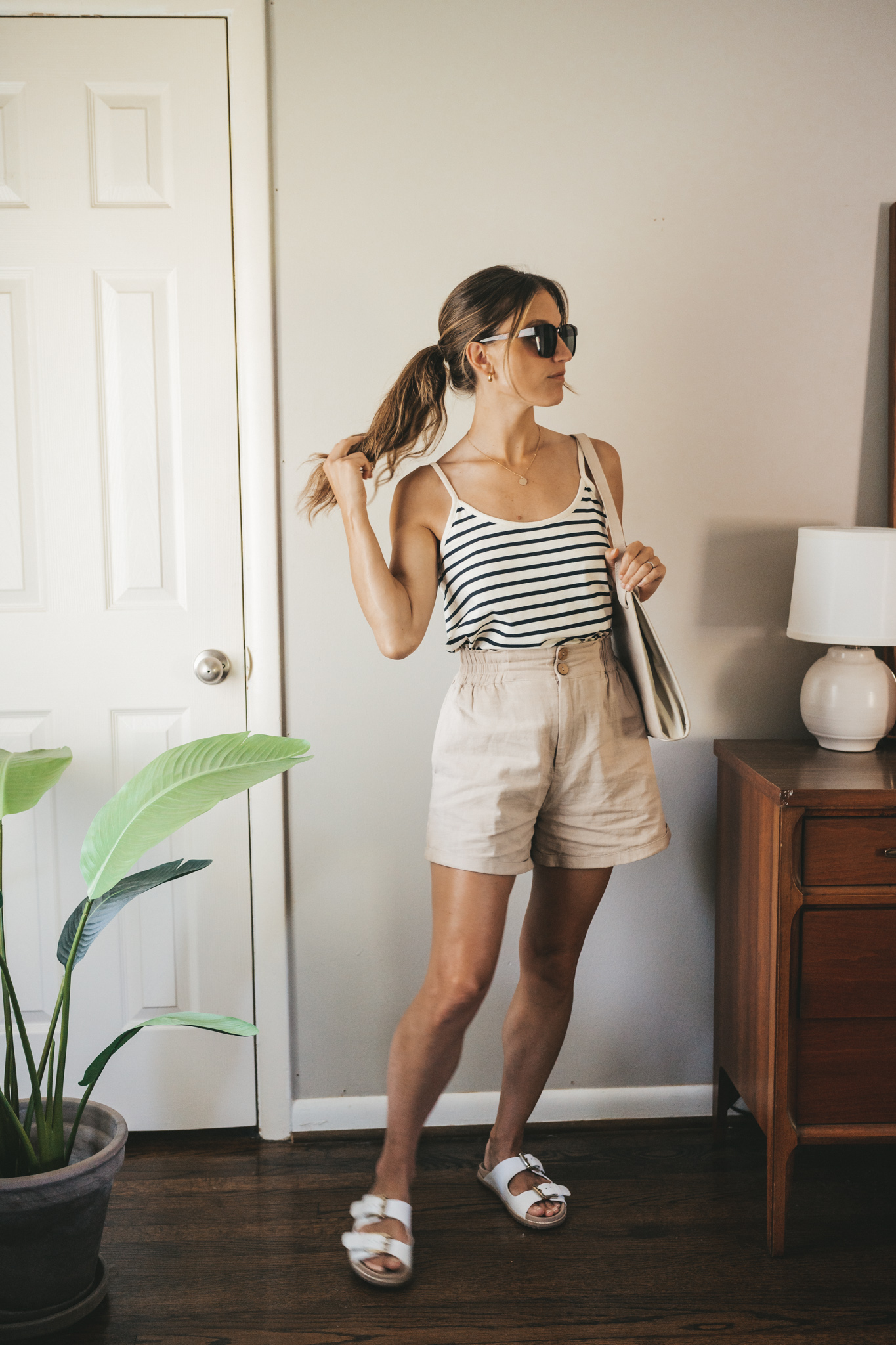 ashley wearing a white and navy striped tank, tan linen shorts, white sandals, a gray shoulder bag, and tortoise shell sunglasses