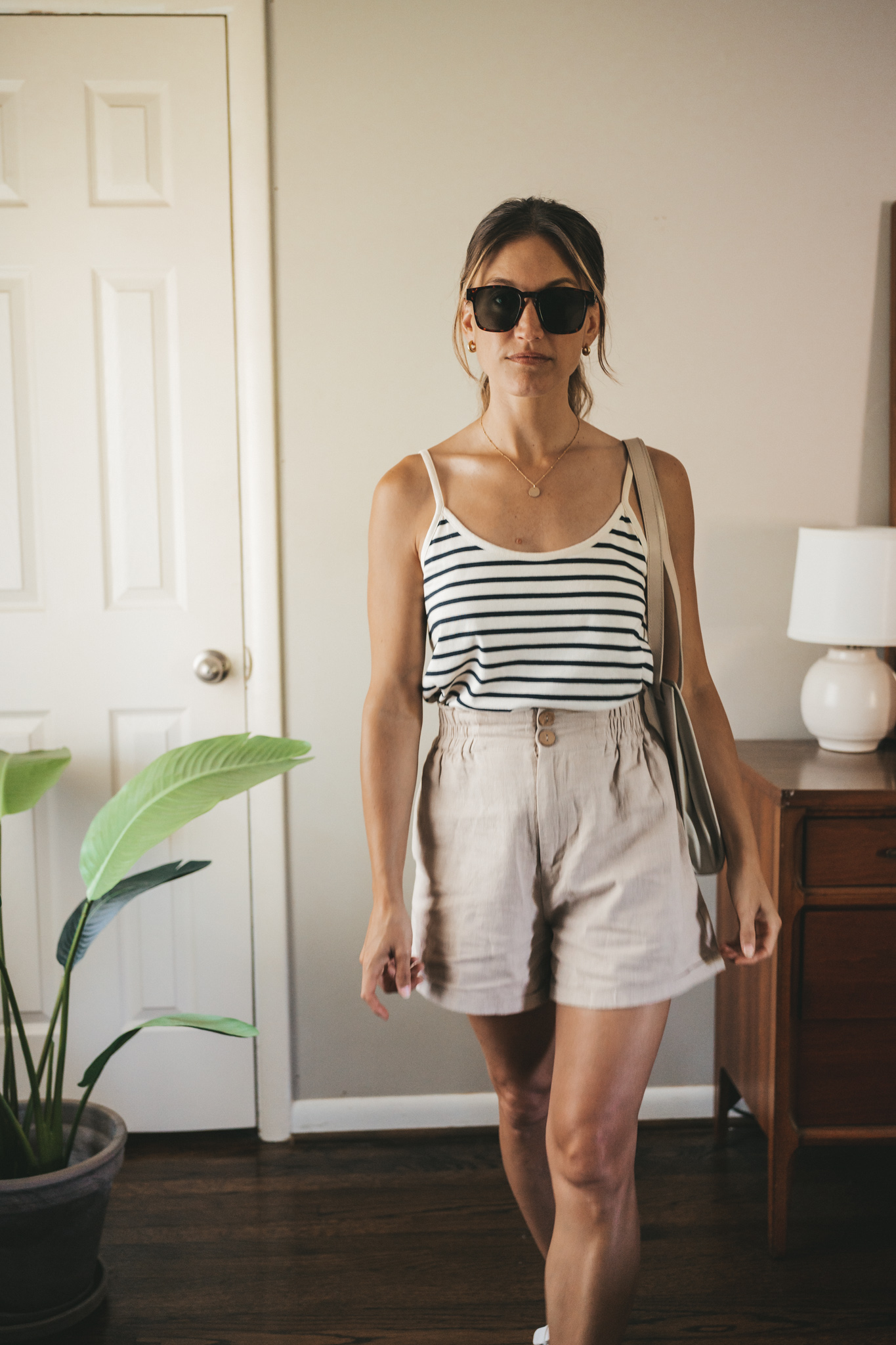 ashley wearing a white and navy striped tank, tan linen shorts, white sandals, a gray shoulder bag, and tortoise shell sunglasses