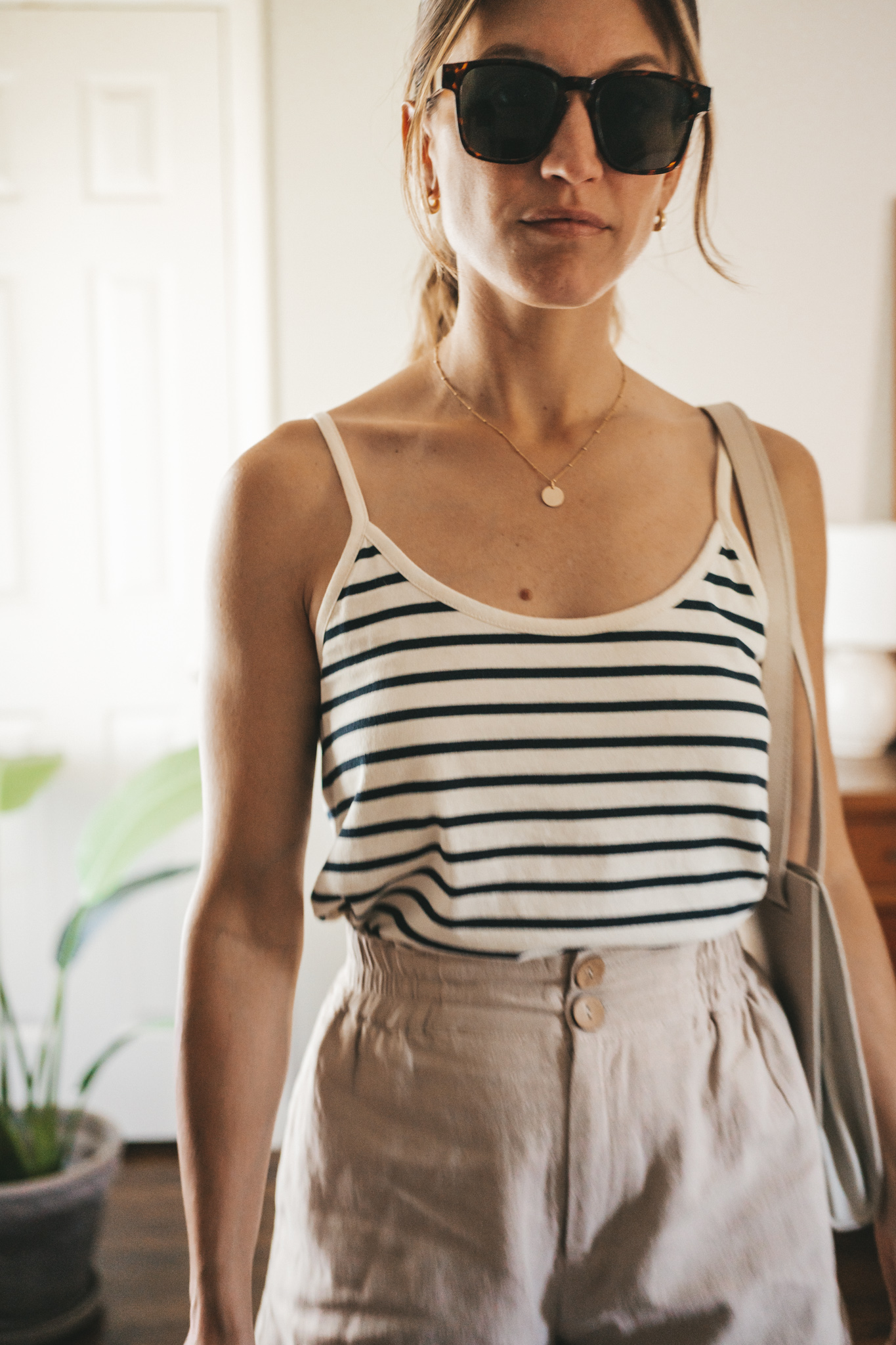 ashley wearing a white and navy striped tank, tan linen shorts, white sandals, a gray shoulder bag, and tortoise shell sunglasses