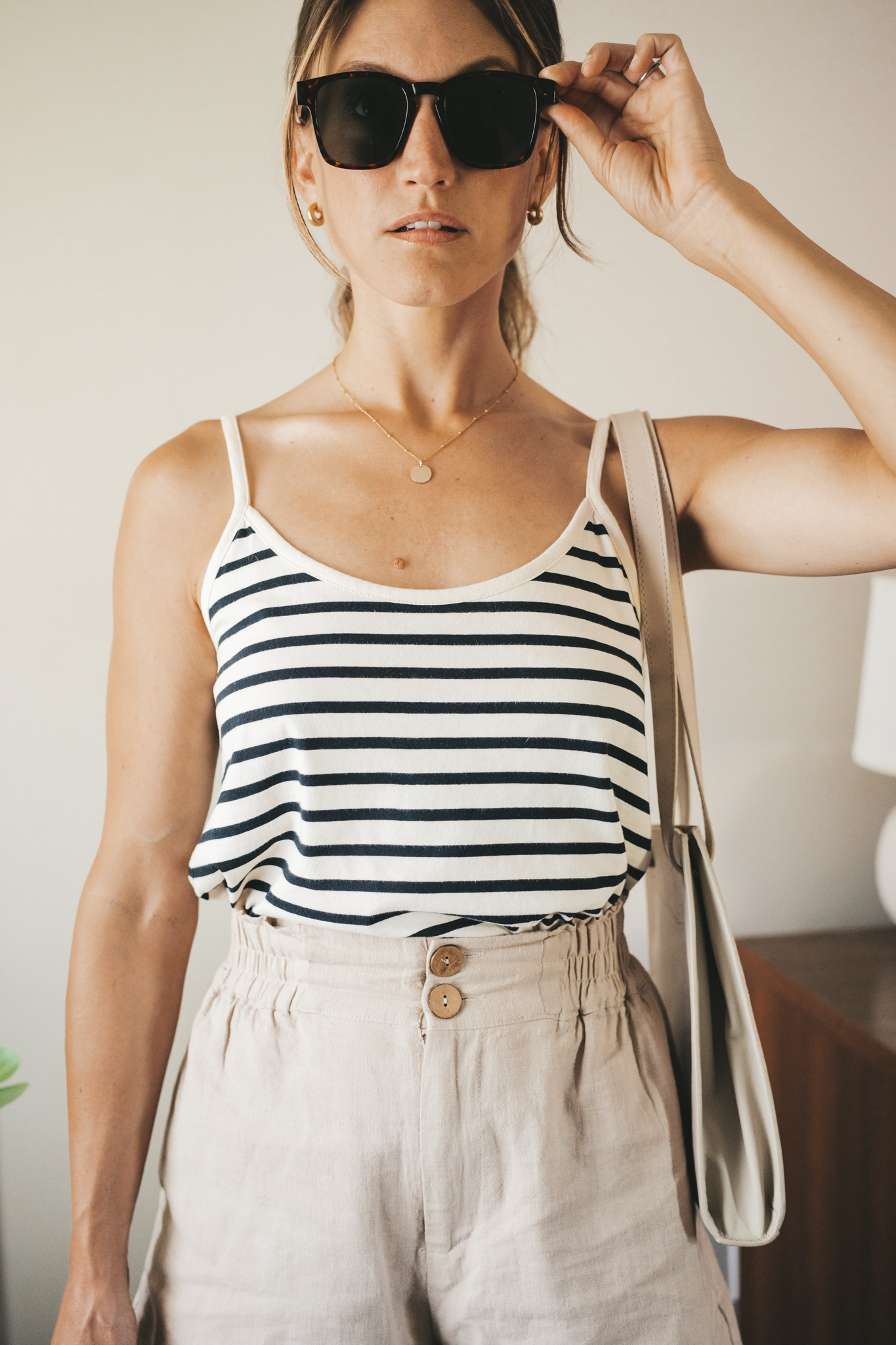 ashley wearing a white and navy striped tank, tan linen shorts, white sandals, a gray shoulder bag, and tortoise shell sunglasses