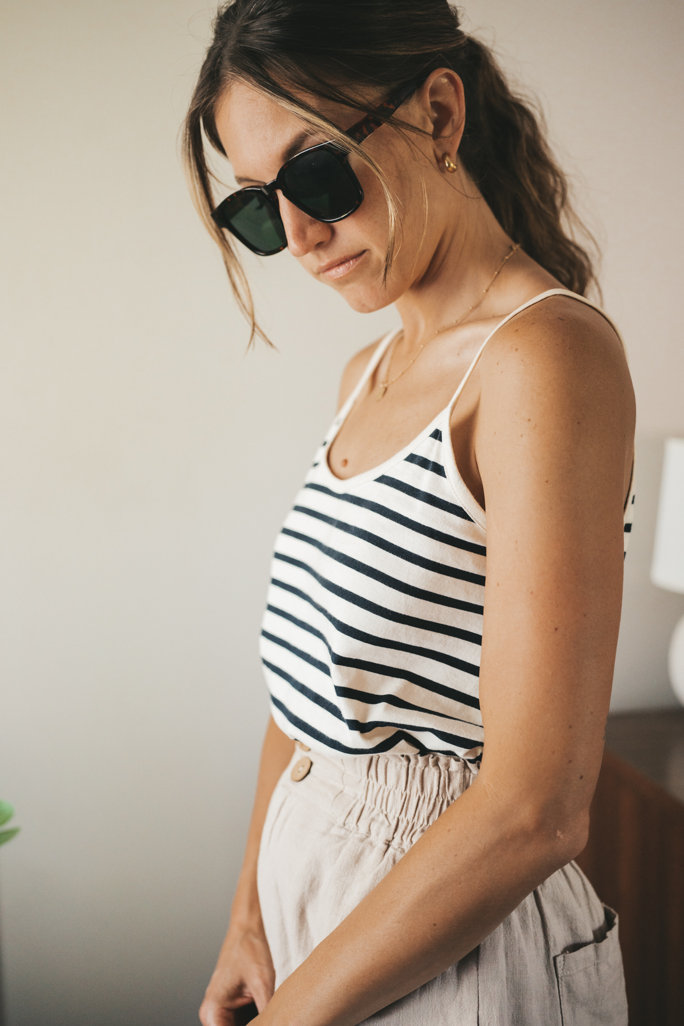 ashley wearing a white and navy striped tank, tan linen shorts, white sandals, a gray shoulder bag, and tortoise shell sunglasses