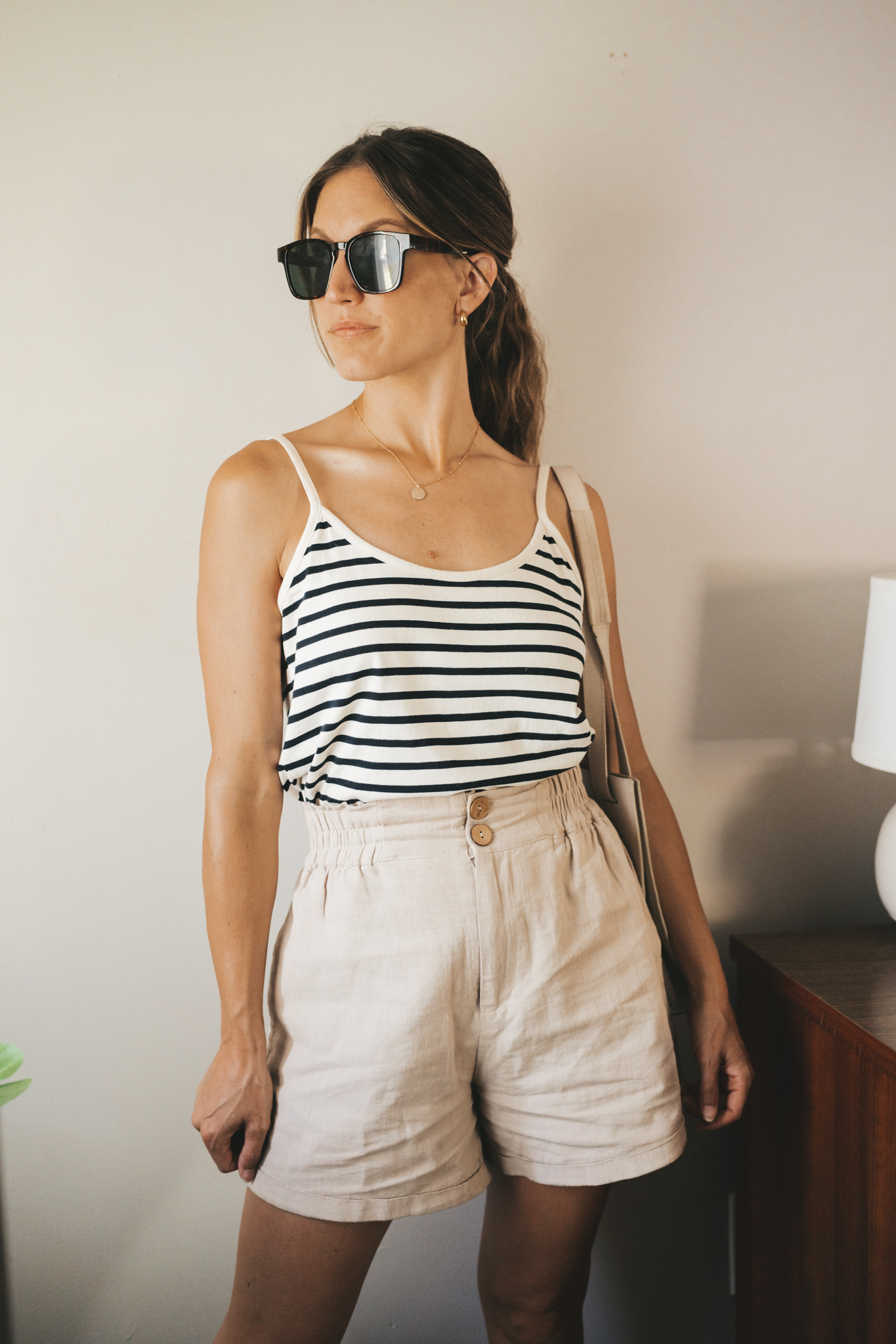 ashley wearing a white and navy striped tank, tan linen shorts, white sandals, a gray shoulder bag, and tortoise shell sunglasses