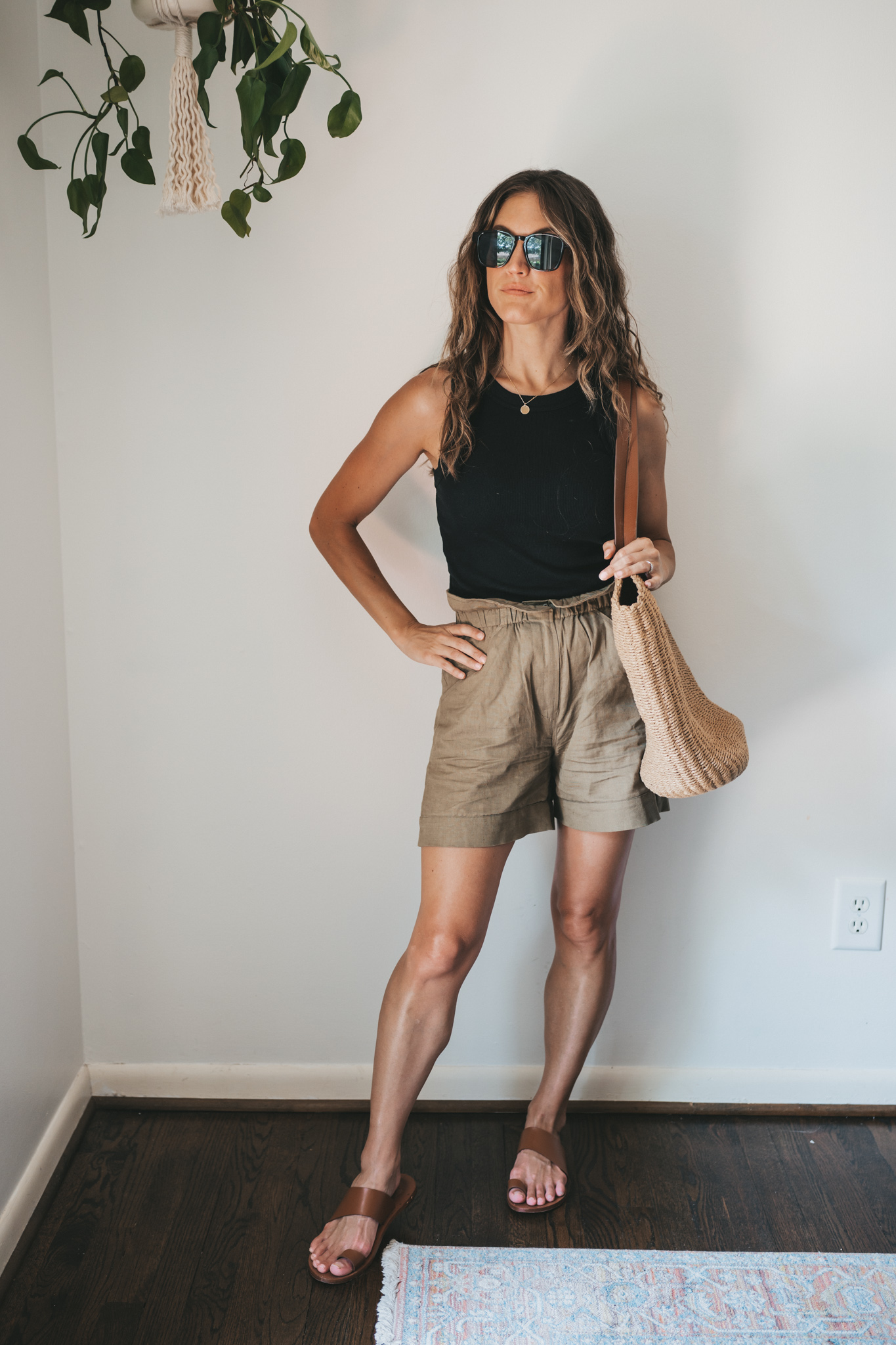 ashley wearing a black ribbed tank, green paperbag shorts, brown leather toe ring sandals, a woven tote bag, and tortoise shell sunglasses