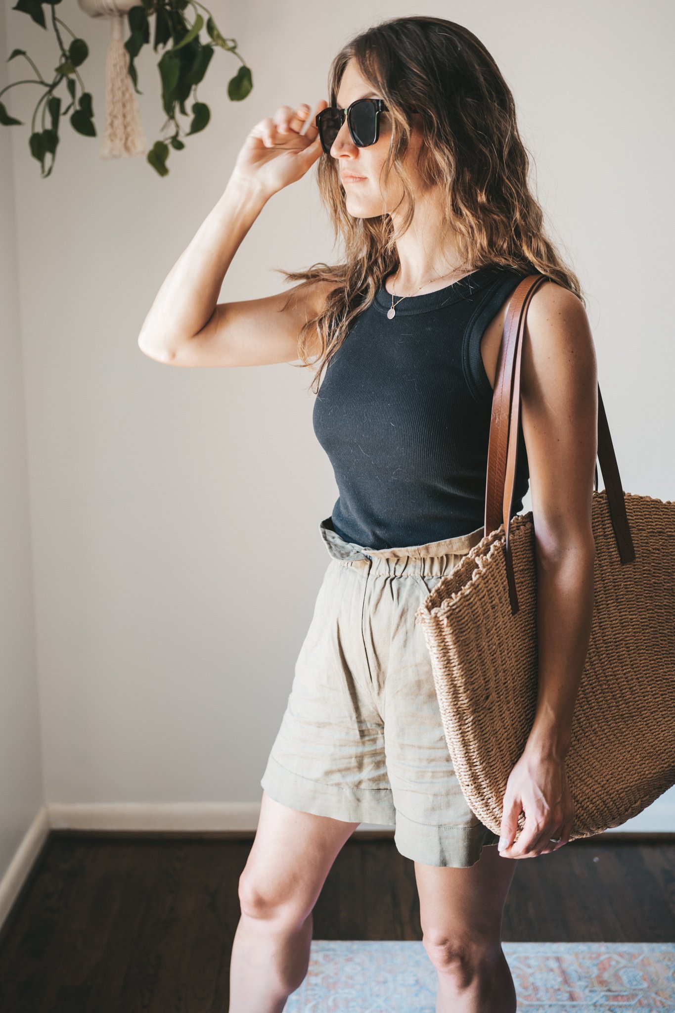 ashley wearing a black ribbed tank, green paperbag shorts, brown leather toe ring sandals, a woven tote bag, and tortoise shell sunglasses