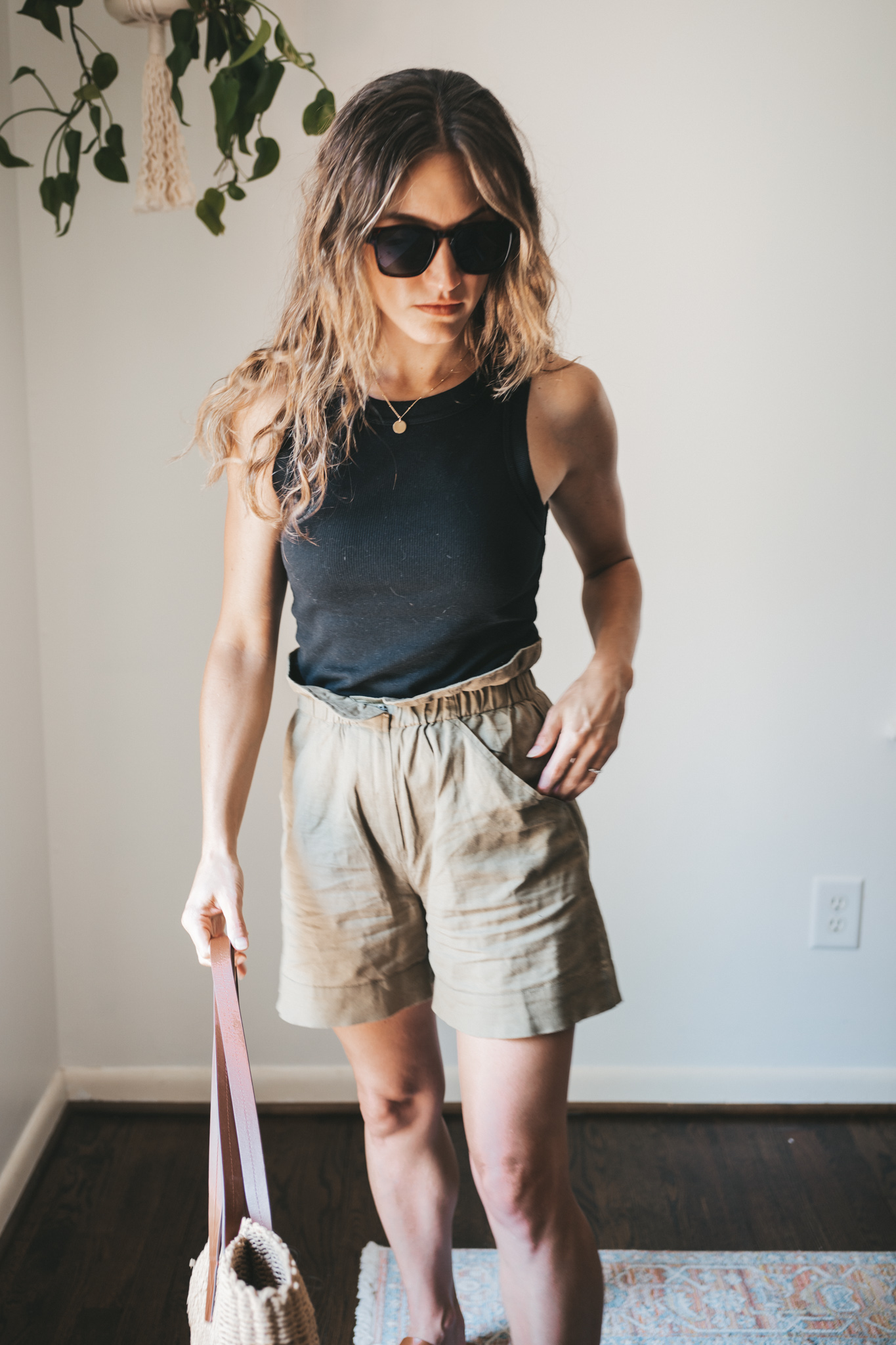 ashley wearing a black ribbed tank, green paperbag shorts, brown leather toe ring sandals, a woven tote bag, and tortoise shell sunglasses