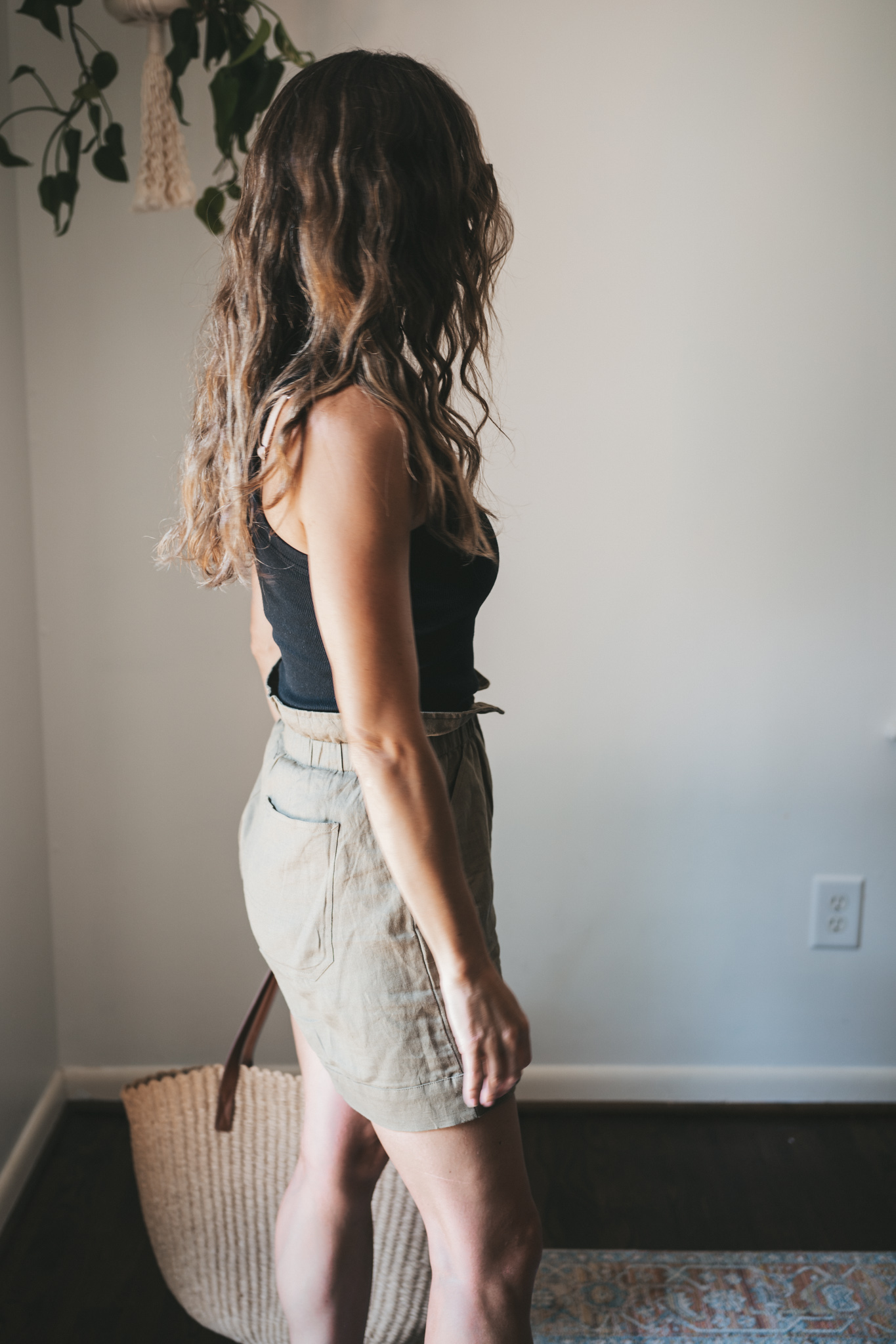 ashley wearing a black ribbed tank, green paperbag shorts, brown leather toe ring sandals, a woven tote bag, and tortoise shell sunglasses