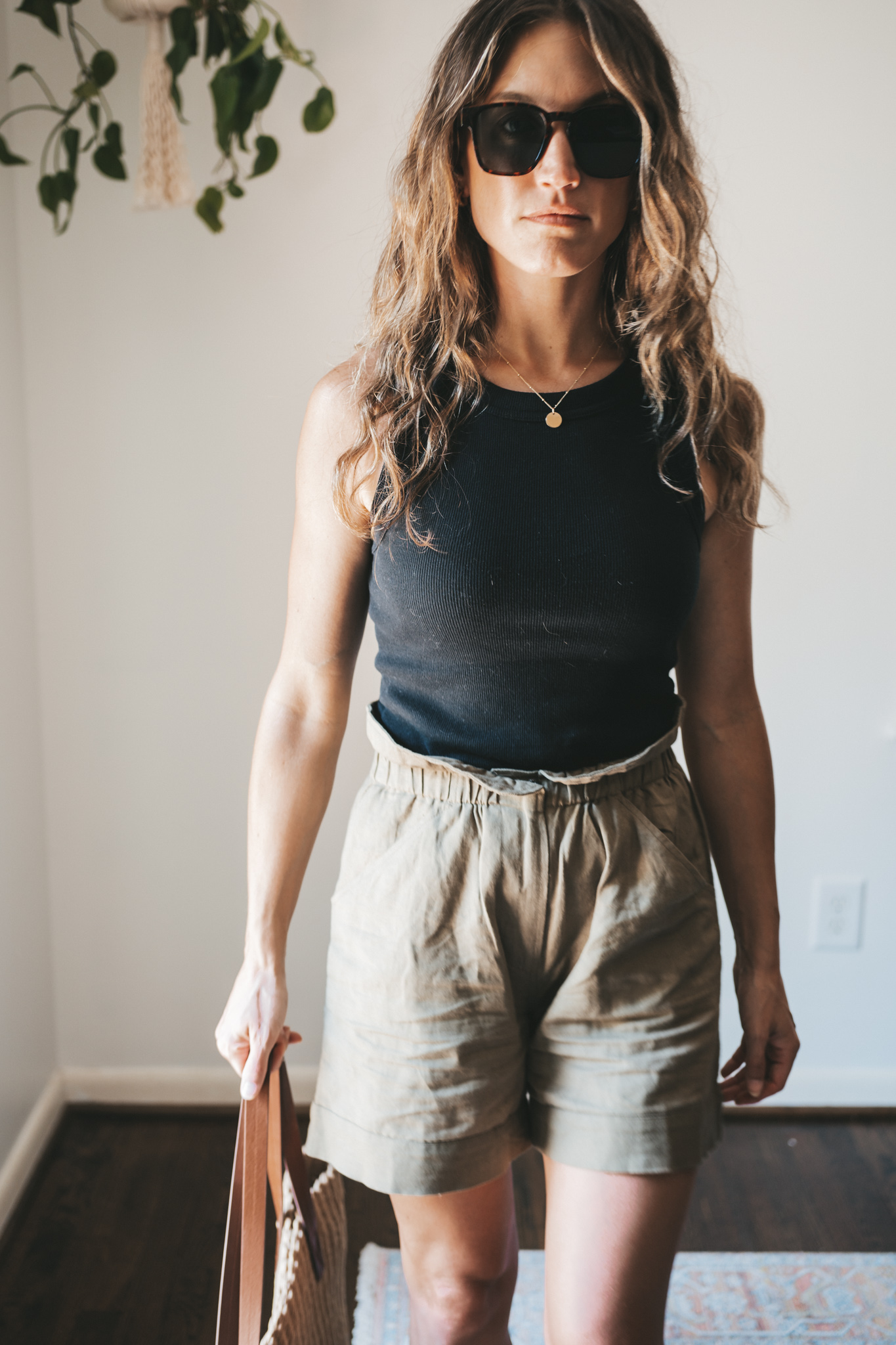 ashley wearing a black ribbed tank, green paperbag shorts, brown leather toe ring sandals, a woven tote bag, and tortoise shell sunglasses