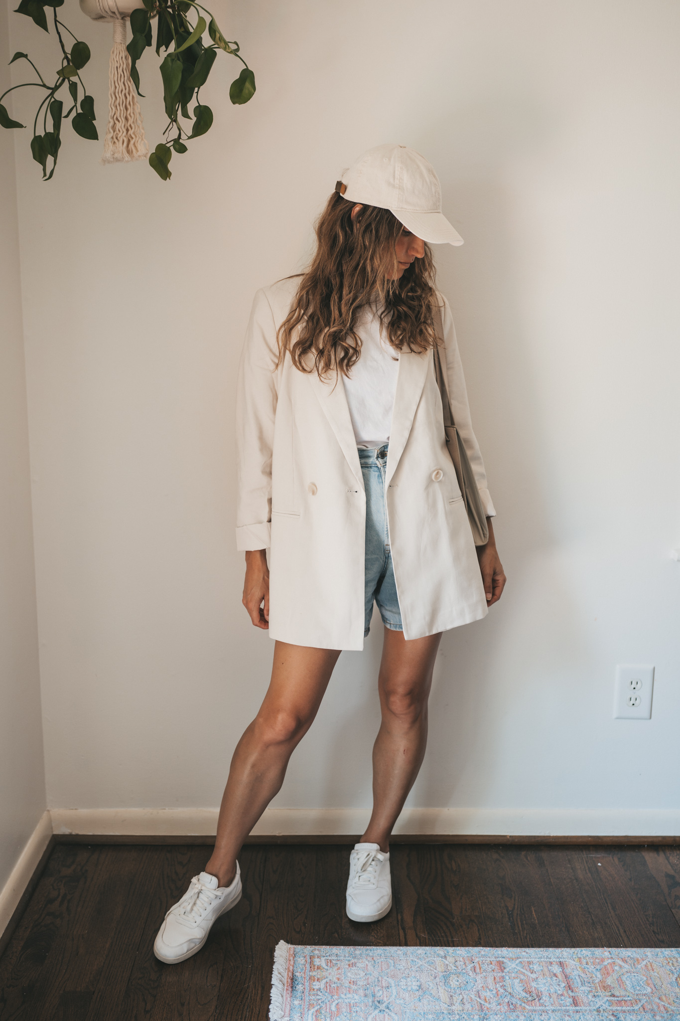 ashley wearing a white tee, beige oversized blazer, light wash long length denim shorts, white sneakers and a beige baseball cap with a gray shoulder bag