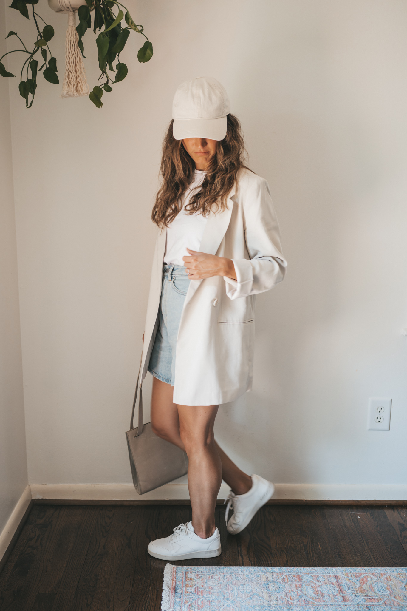 ashley wearing a white tee, beige oversized blazer, light wash long length denim shorts, white sneakers and a beige baseball cap with a gray shoulder bag