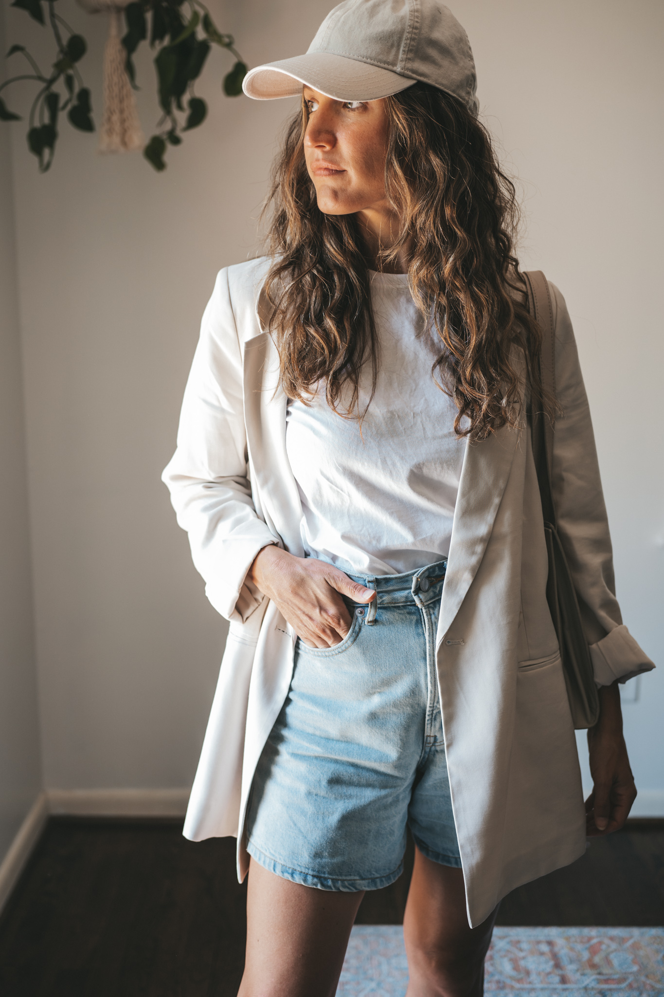 ashley wearing a white tee, beige oversized blazer, light wash long length denim shorts, white sneakers and a beige baseball cap with a gray shoulder bag