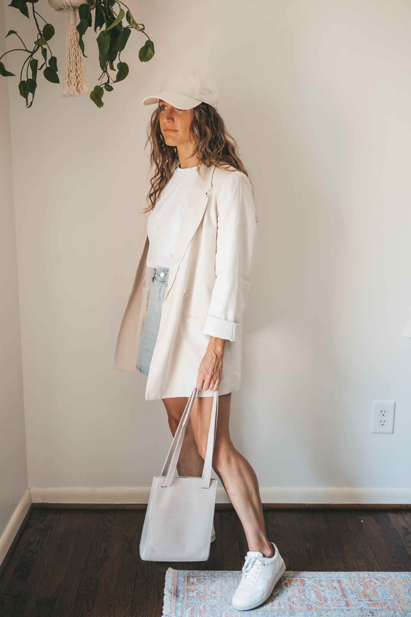 ashley wearing a white tee, beige oversized blazer, light wash long length denim shorts, white sneakers and a beige baseball cap with a gray shoulder bag