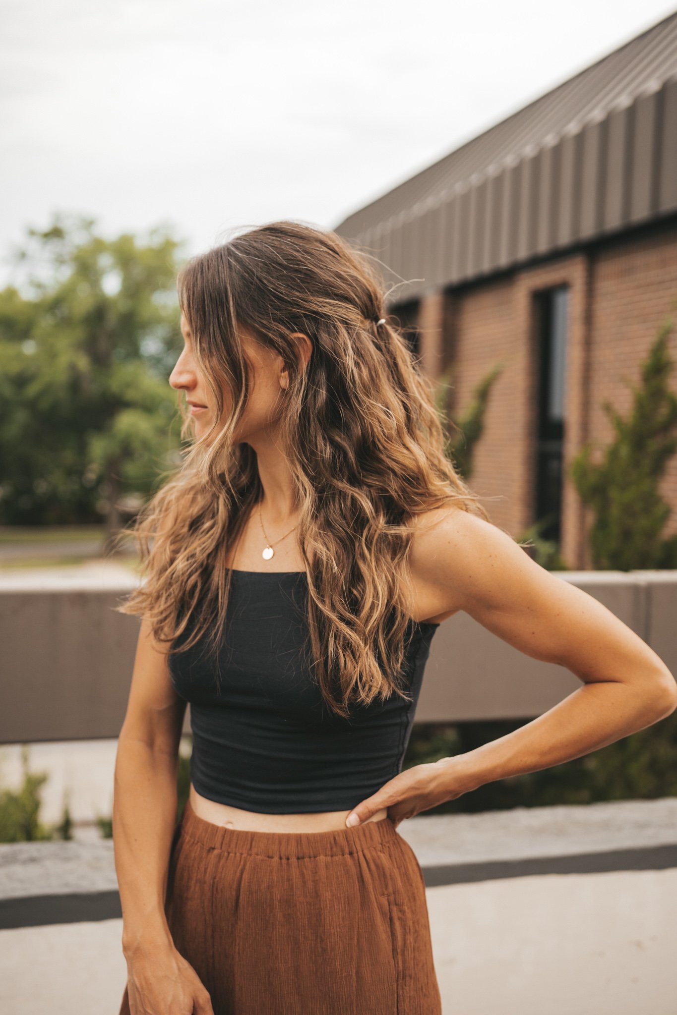ashley wearing a black cropped cami tank, brown tapered pants, black strappy sandals, and a woven tote bag standing outside next to a brown building