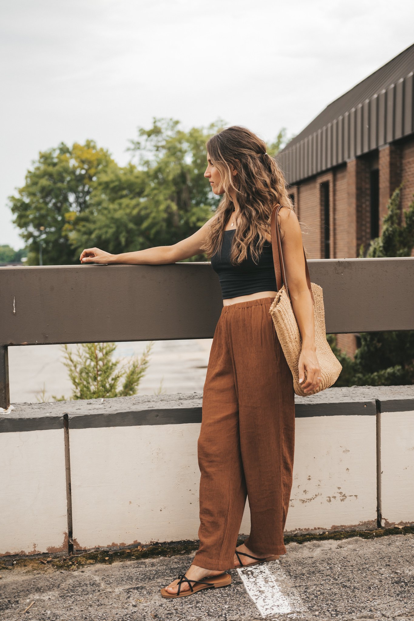 ashley wearing a black cropped cami tank, brown tapered pants, black strappy sandals, and a woven tote bag standing outside next to a brown building