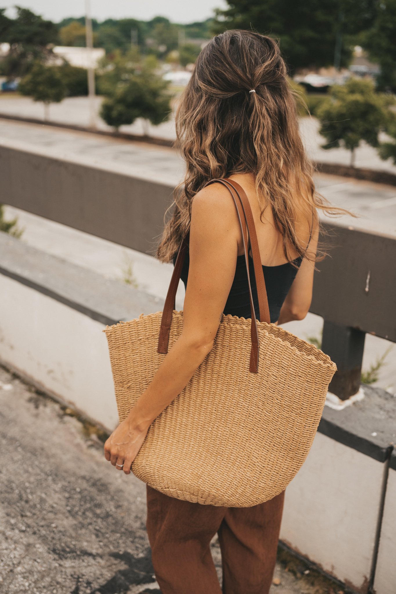 ashley wearing a black cropped cami tank, brown tapered pants, black strappy sandals, and a woven tote bag standing outside next to a brown building