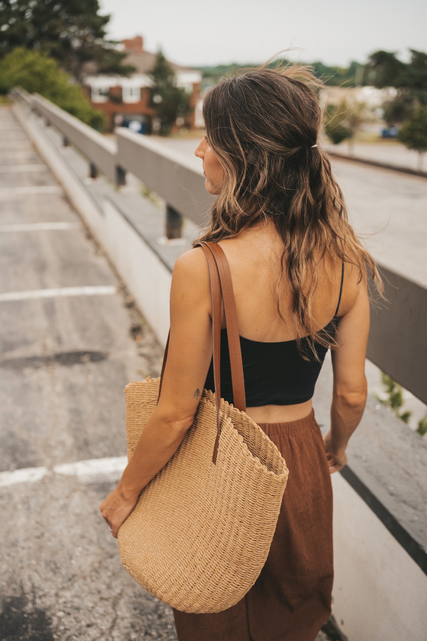 ashley wearing a black cropped cami tank, brown tapered pants, black strappy sandals, and a woven tote bag standing outside next to a brown building