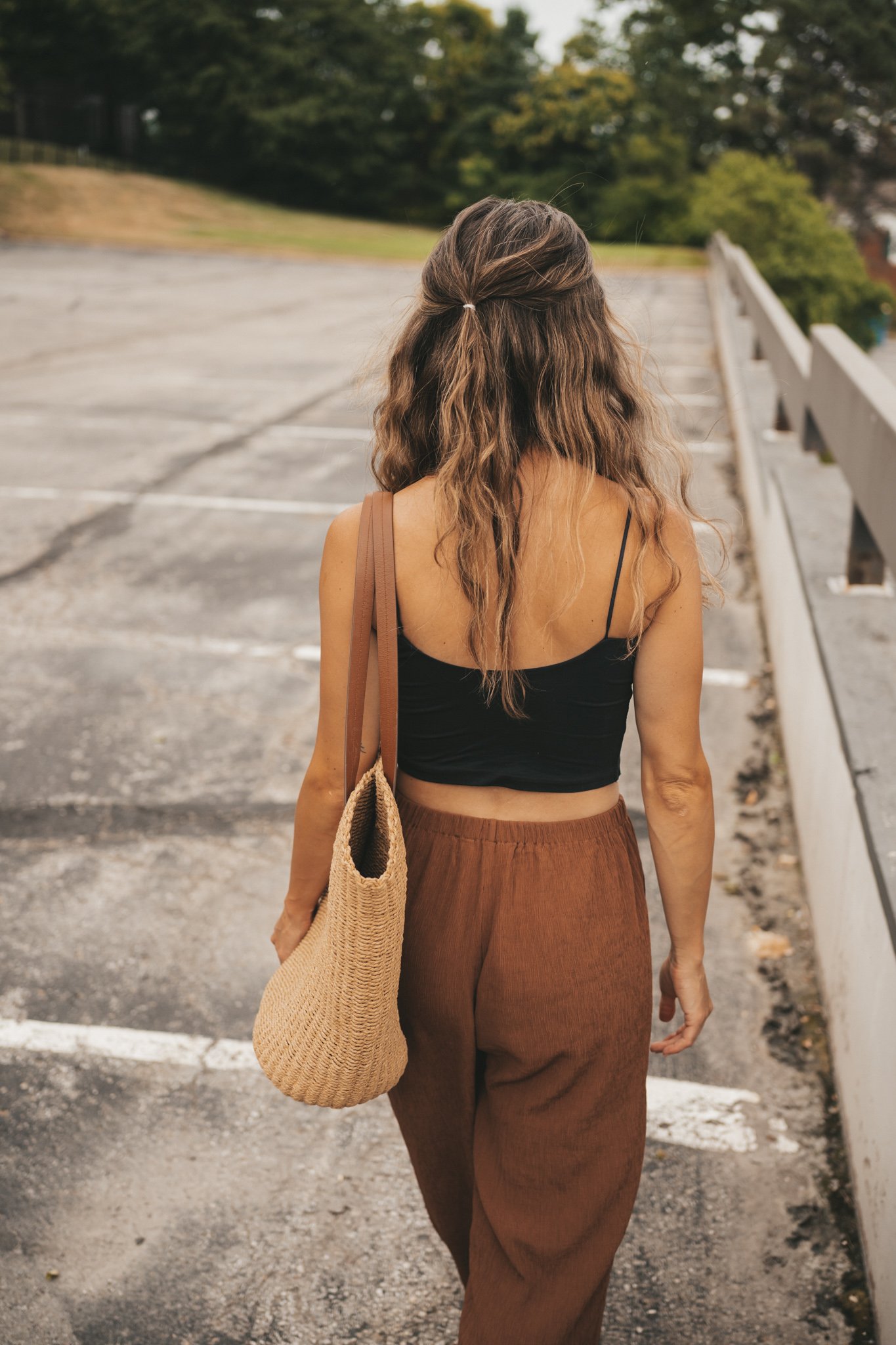 ashley wearing a black cropped cami tank, brown tapered pants, black strappy sandals, and a woven tote bag standing outside next to a brown building