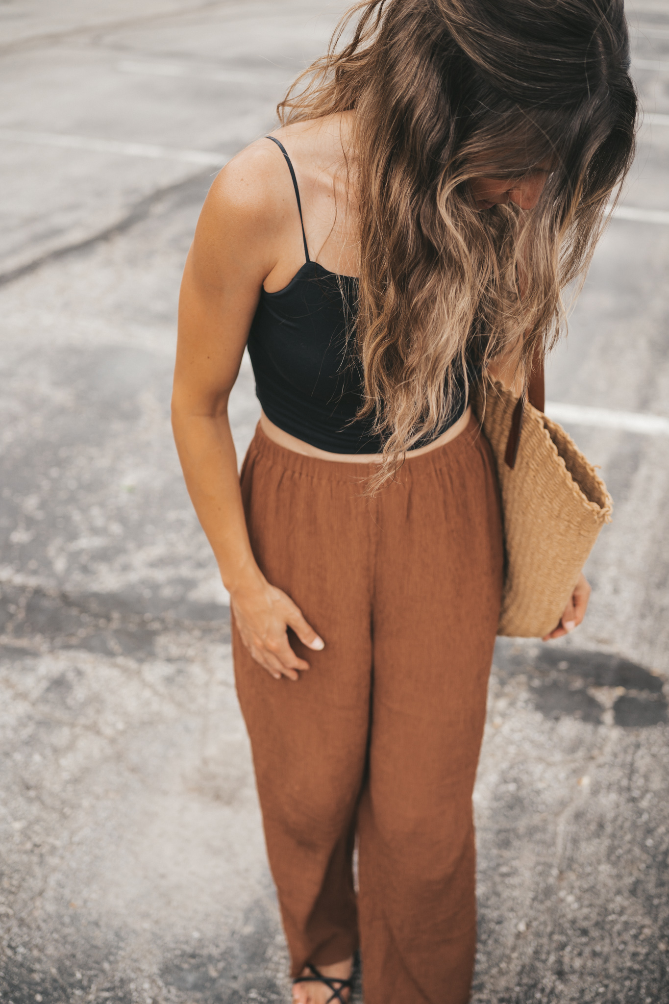 ashley wearing a black cropped cami tank, brown tapered pants, black strappy sandals, and a woven tote bag standing outside next to a brown building