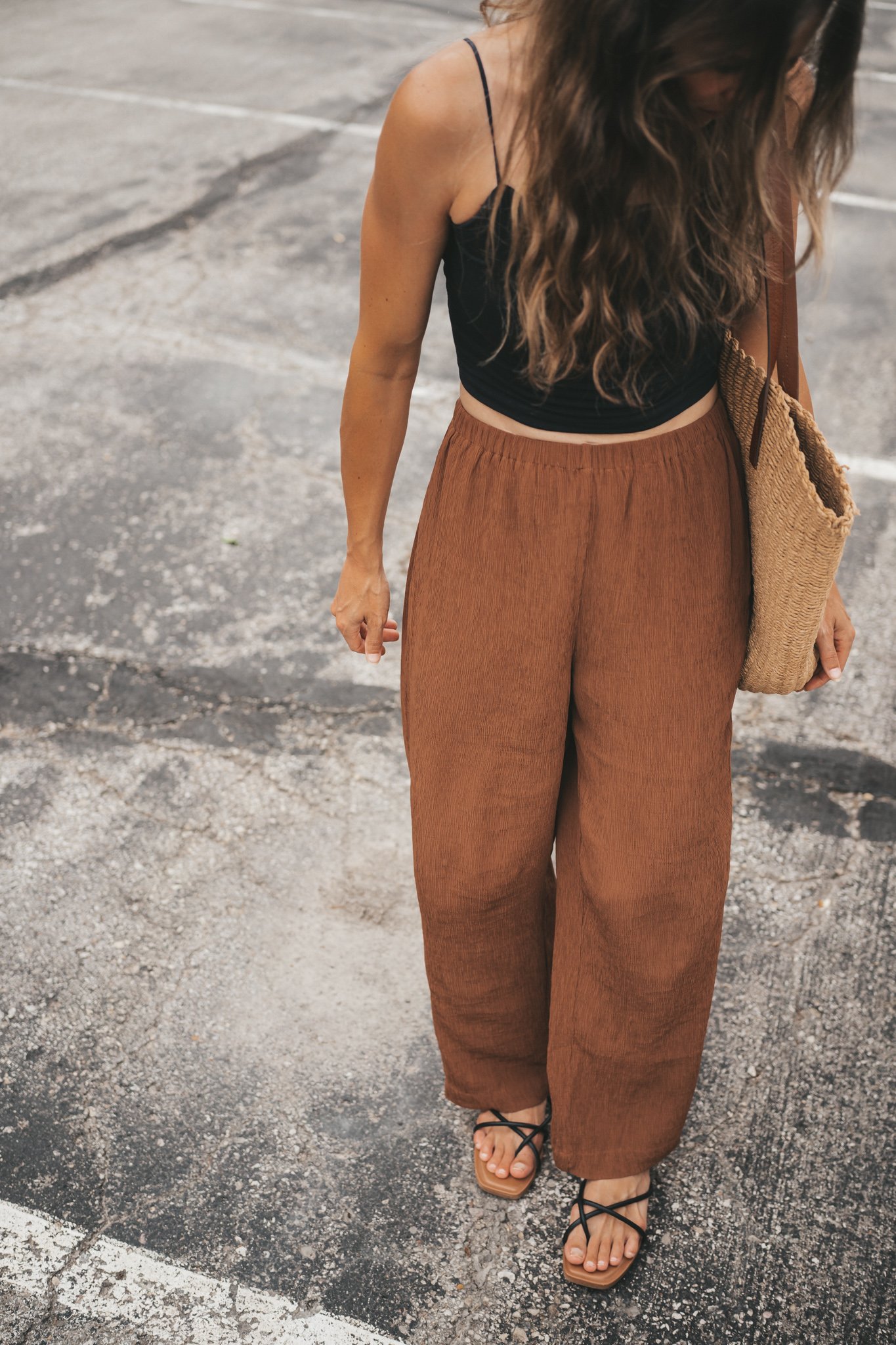 ashley wearing a black cropped cami tank, brown tapered pants, black strappy sandals, and a woven tote bag standing outside next to a brown building