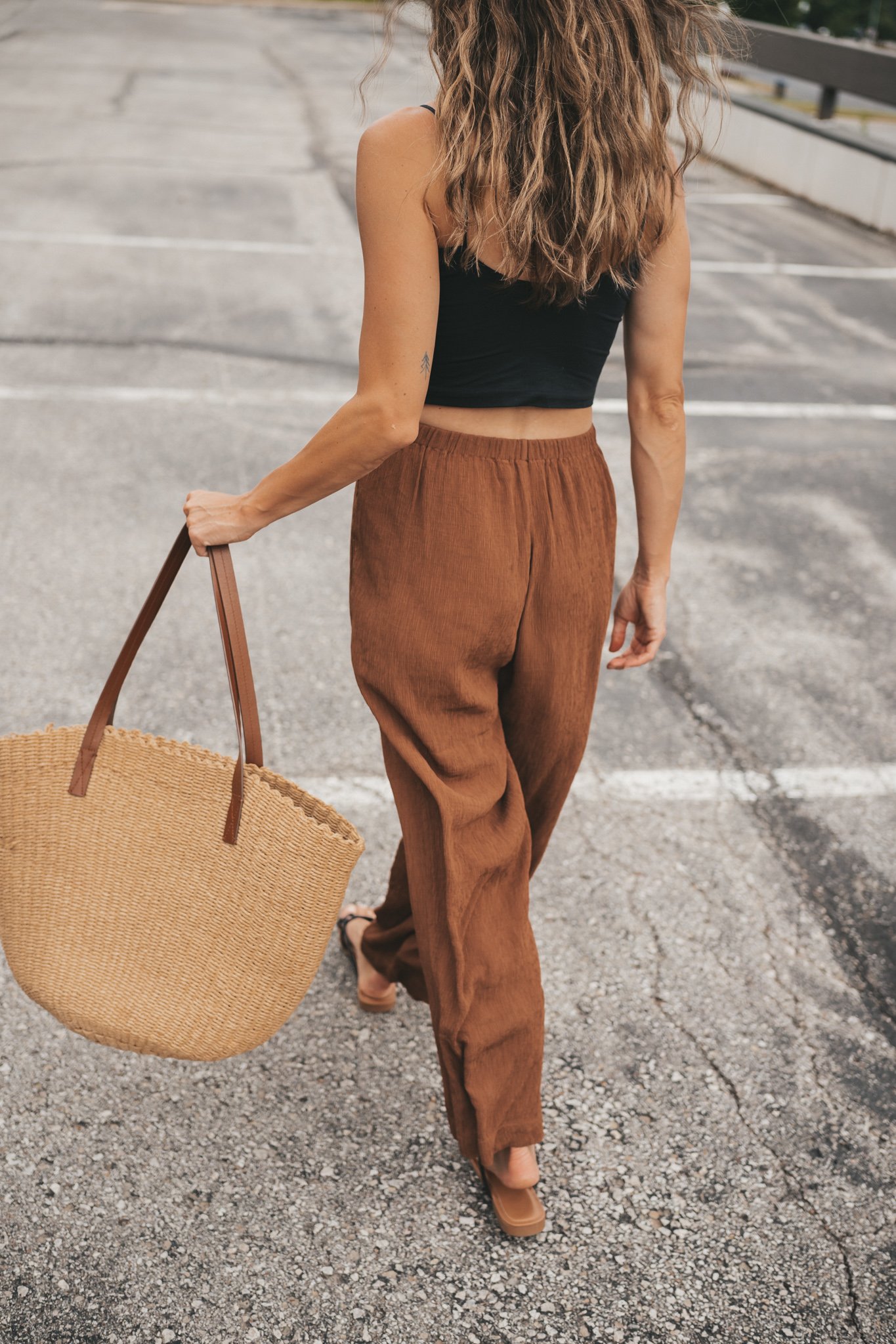 ashley wearing a black cropped cami tank, brown tapered pants, black strappy sandals, and a woven tote bag standing outside next to a brown building
