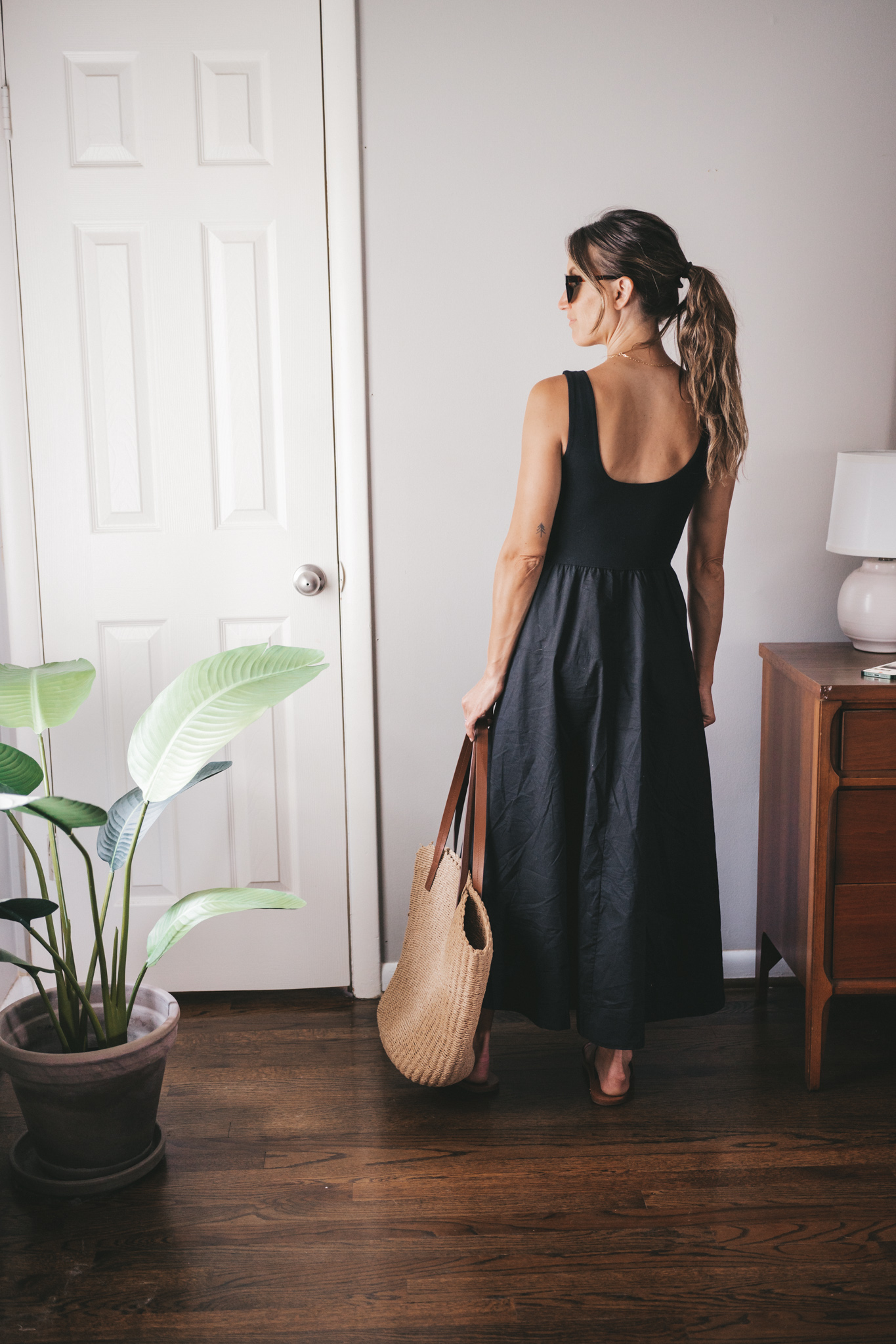 ashley wearing a black A-Line dress, brown leather sandals and a tan woven tote bag