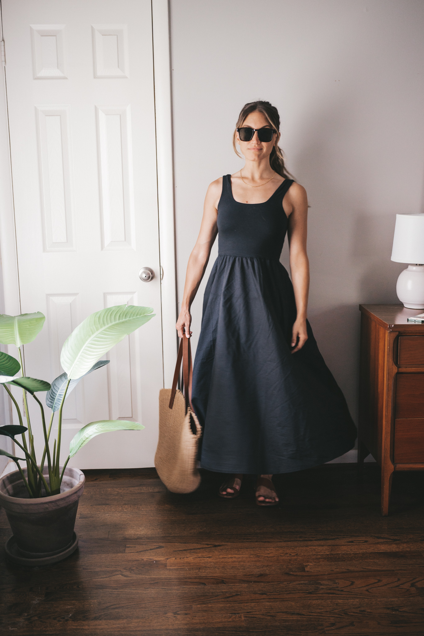 ashley wearing a black A-Line dress, brown leather sandals and a tan woven tote bag