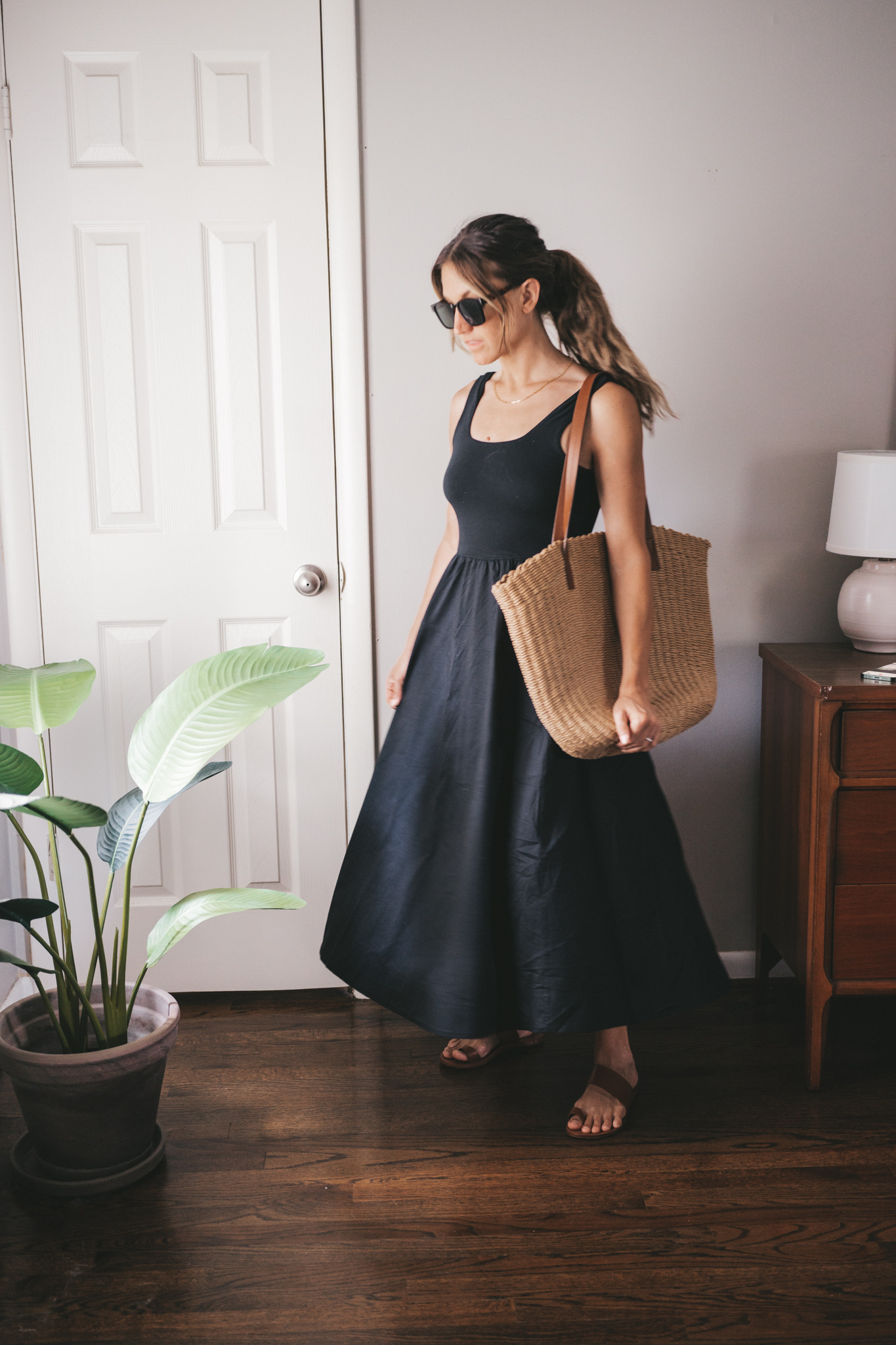 ashley wearing a black A-Line dress, brown leather sandals and a tan woven tote bag