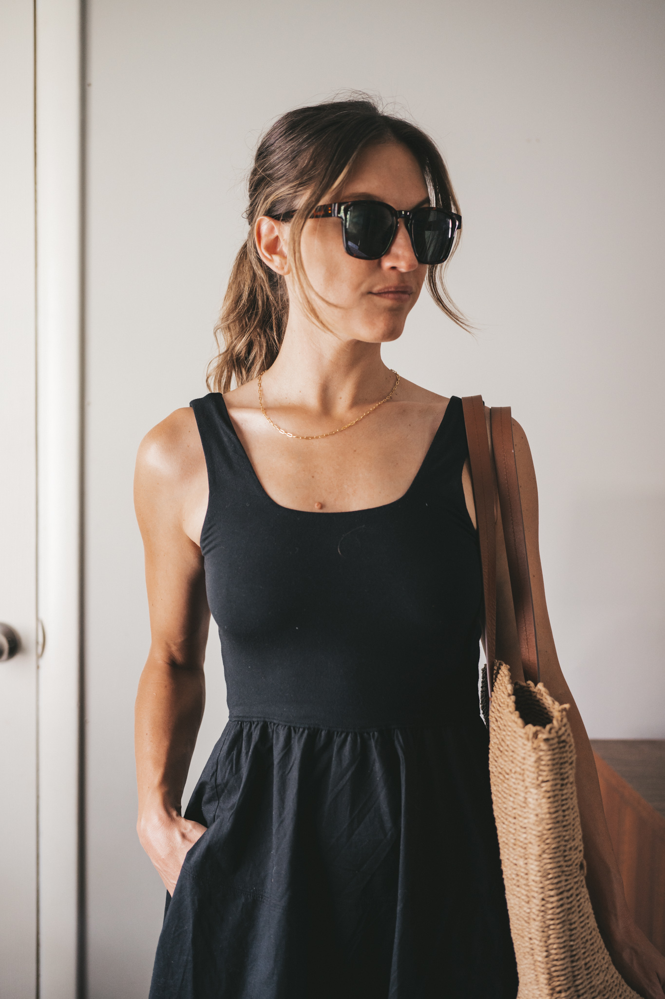ashley wearing a black A-Line dress, brown leather sandals and a tan woven tote bag