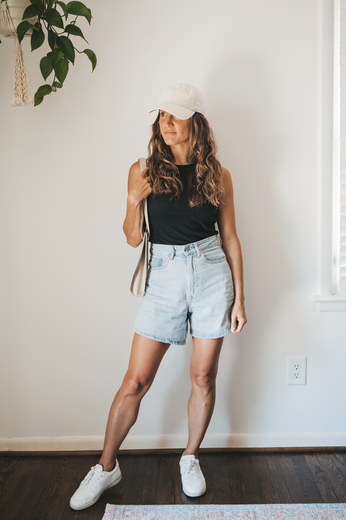 ashley wearing a black ribbed tank, light wash denim shorts, white sneakers, a beige baseball cap, and a gray leather shoulder bag