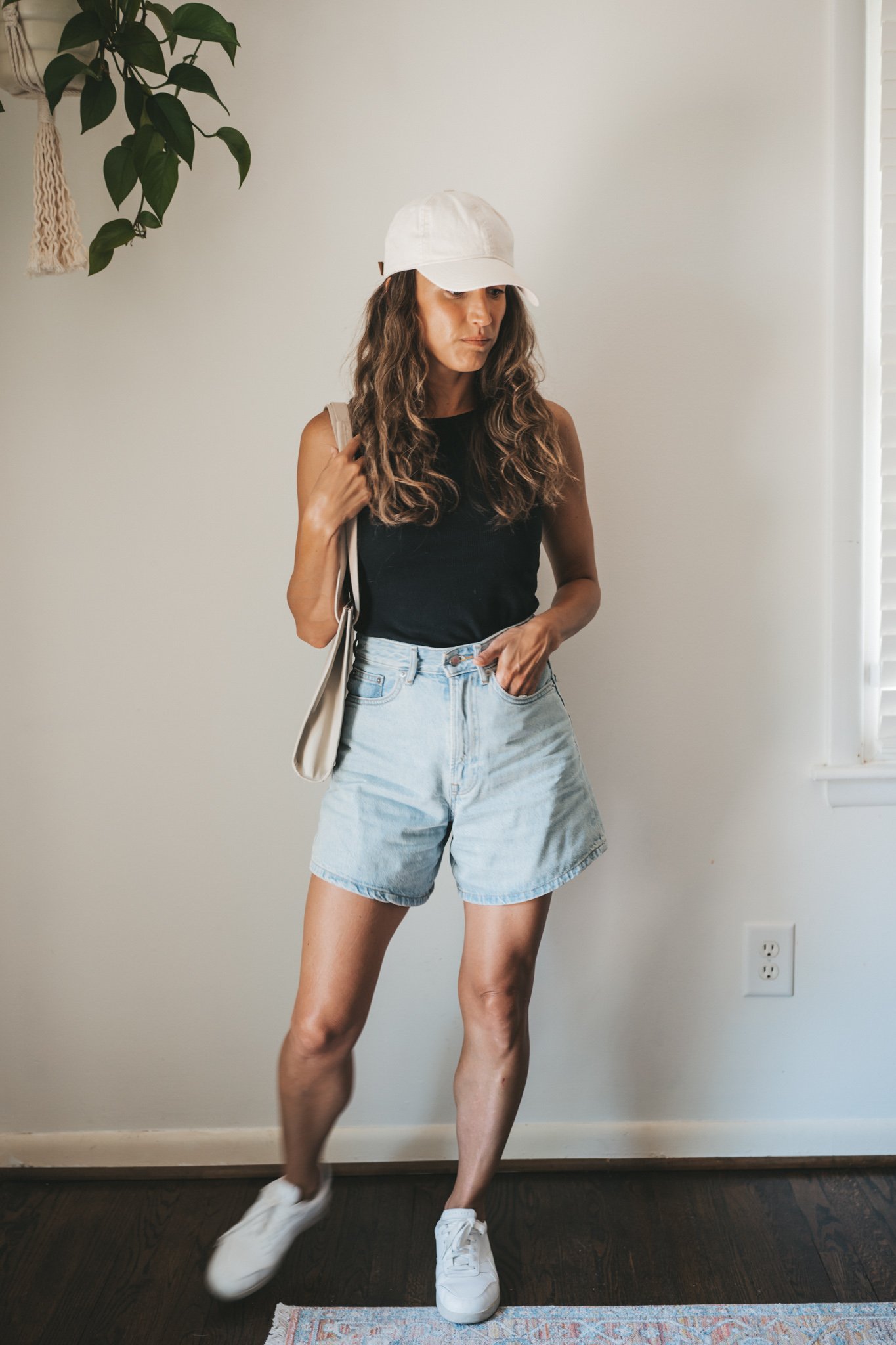 ashley wearing a black ribbed tank, light wash denim shorts, white sneakers, a beige baseball cap, and a gray leather shoulder bag