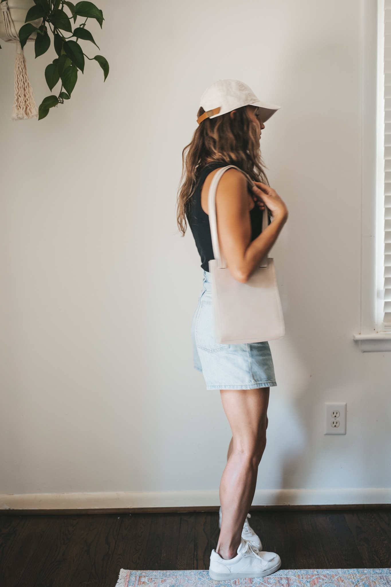 ashley wearing a black ribbed tank, light wash denim shorts, white sneakers, a beige baseball cap, and a gray leather shoulder bag