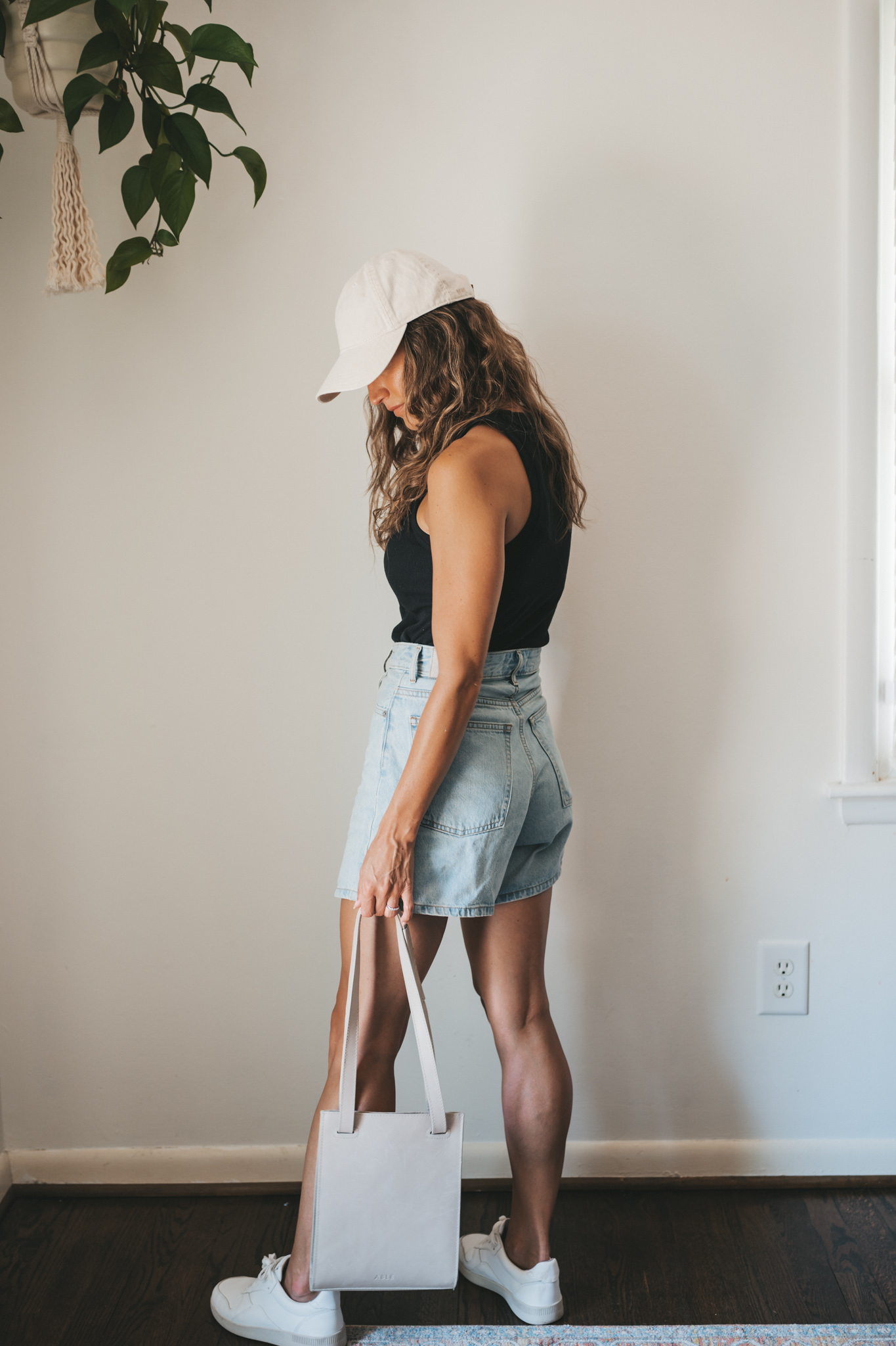 ashley wearing a black ribbed tank, light wash denim shorts, white sneakers, a beige baseball cap, and a gray leather shoulder bag