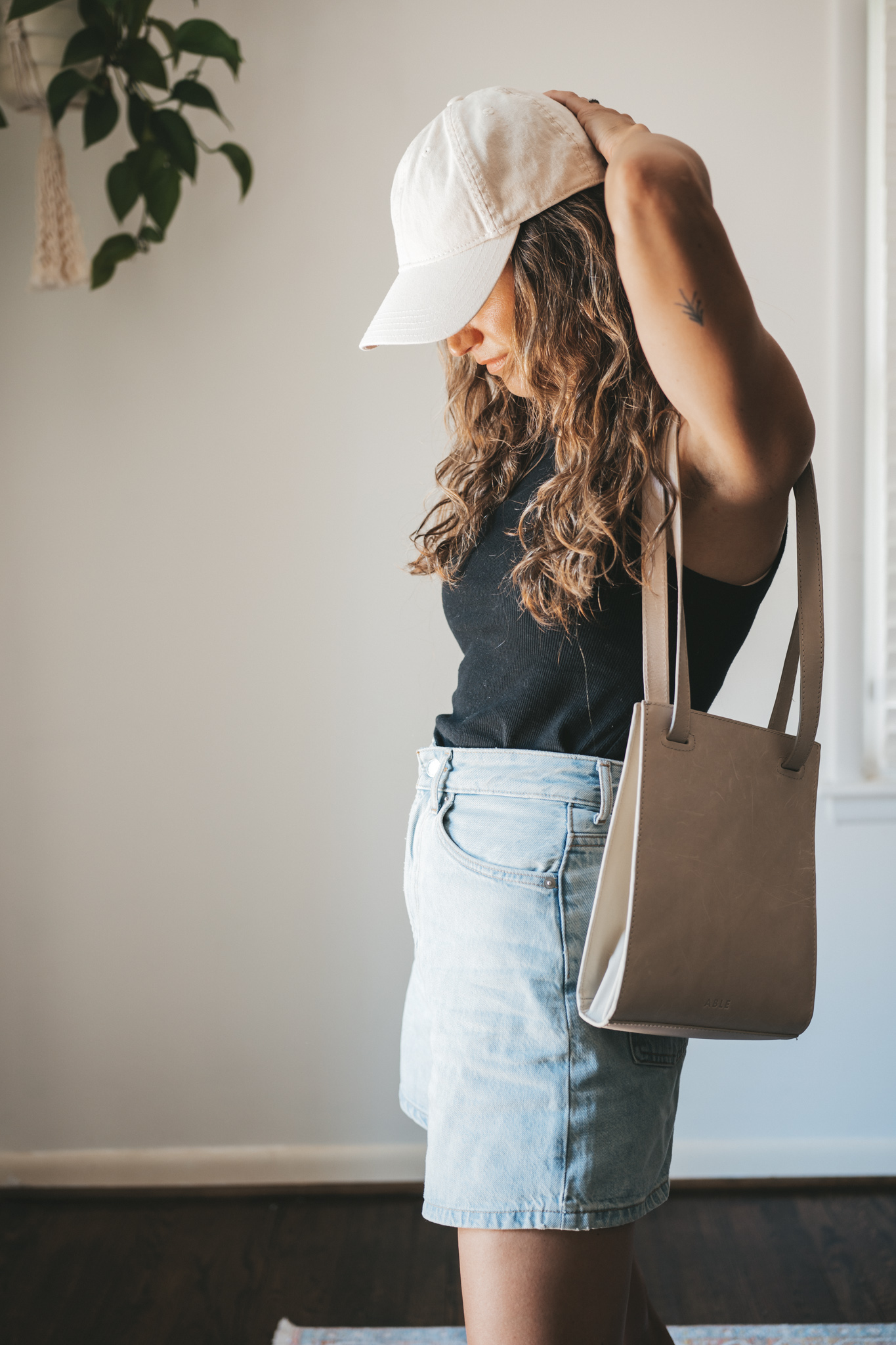 ashley wearing a black ribbed tank, light wash denim shorts, white sneakers, a beige baseball cap, and a gray leather shoulder bag