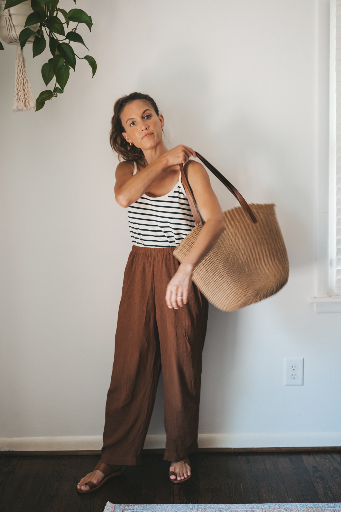 ashley wearing a white and navy striped tank, chocolate brown tapered loose pants, brown leather sandals, and a woven tote bag