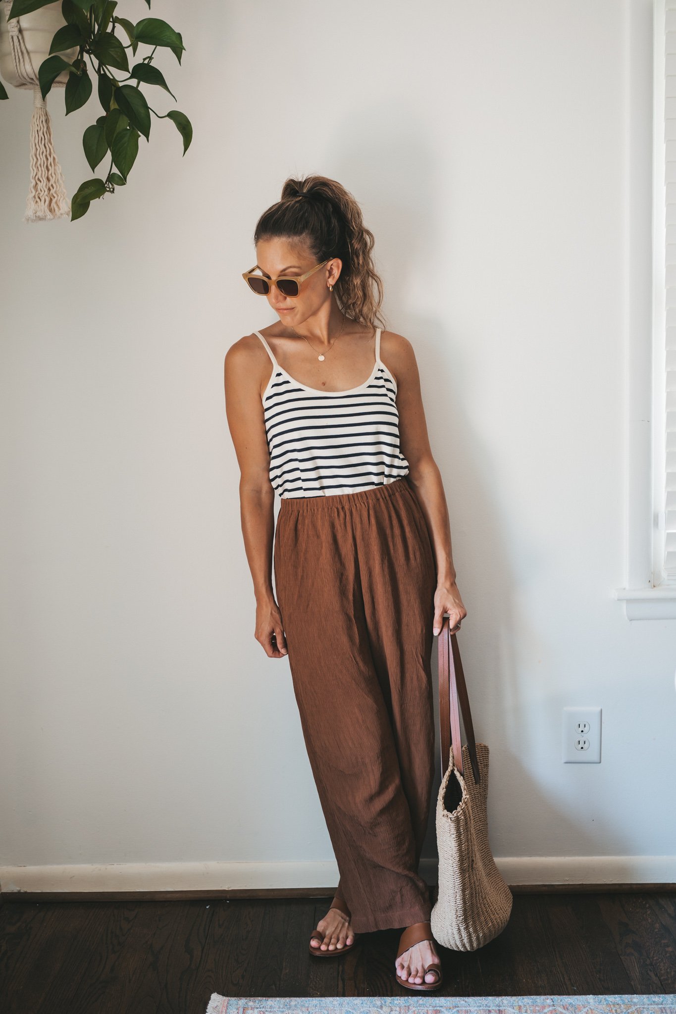ashley wearing a white and navy striped tank, chocolate brown tapered loose pants, brown leather sandals, and a woven tote bag