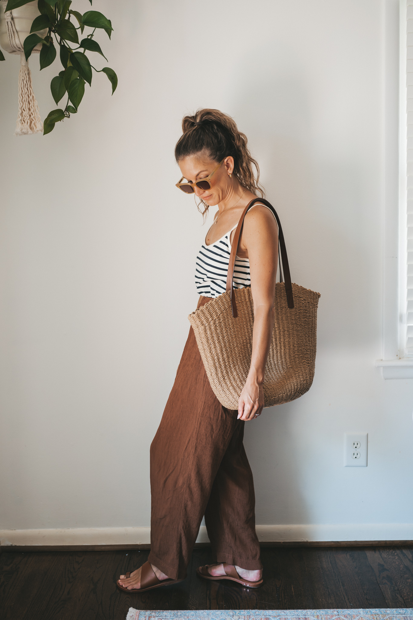 ashley wearing a white and navy striped tank, chocolate brown tapered loose pants, brown leather sandals, and a woven tote bag