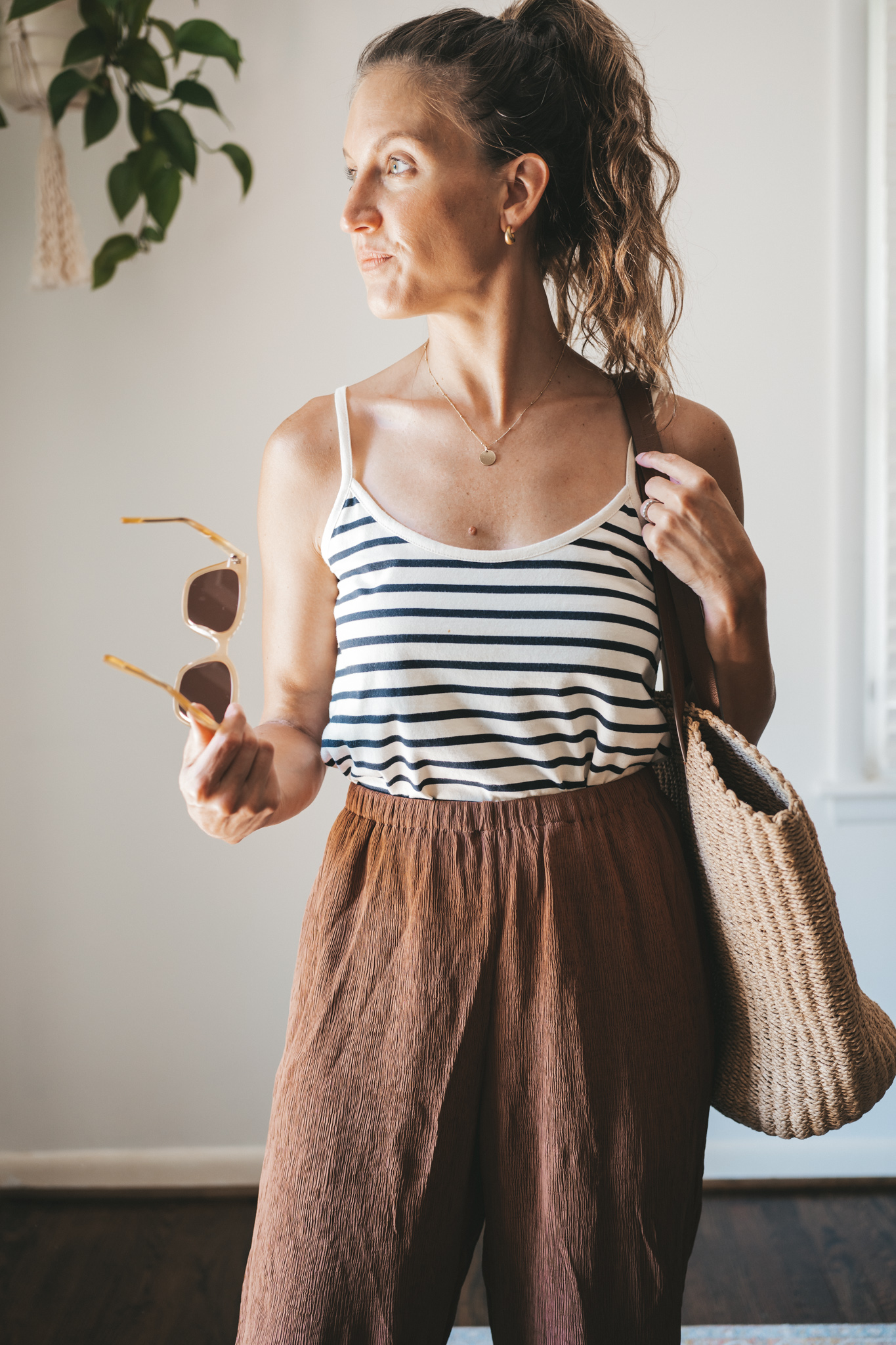 ashley wearing a white and navy striped tank, chocolate brown tapered loose pants, brown leather sandals, and a woven tote bag
