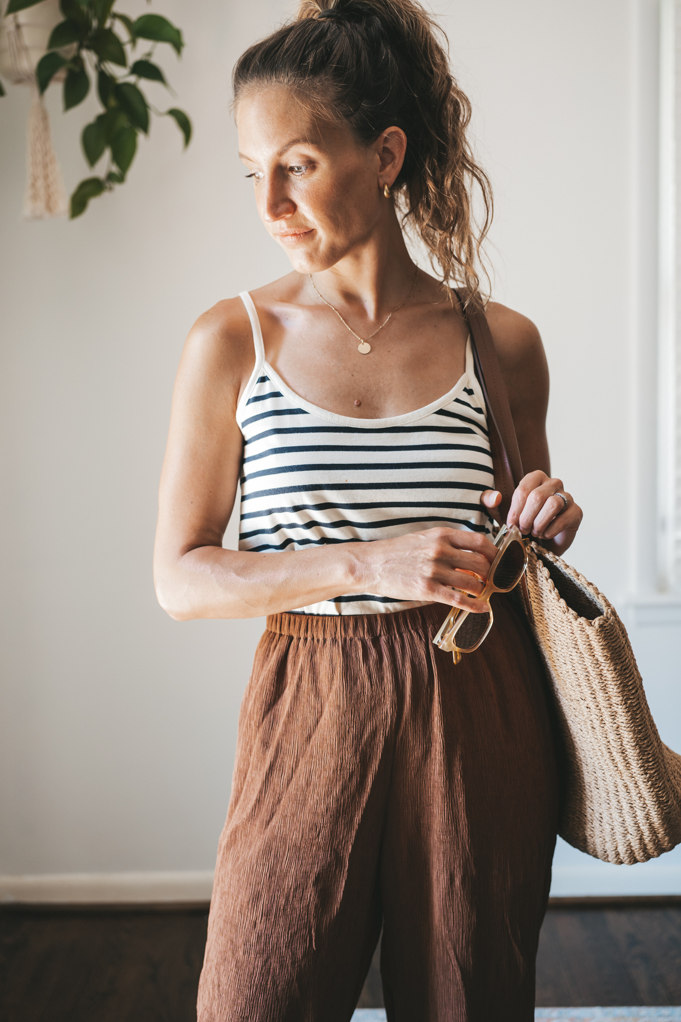 ashley wearing a white and navy striped tank, chocolate brown tapered loose pants, brown leather sandals, and a woven tote bag