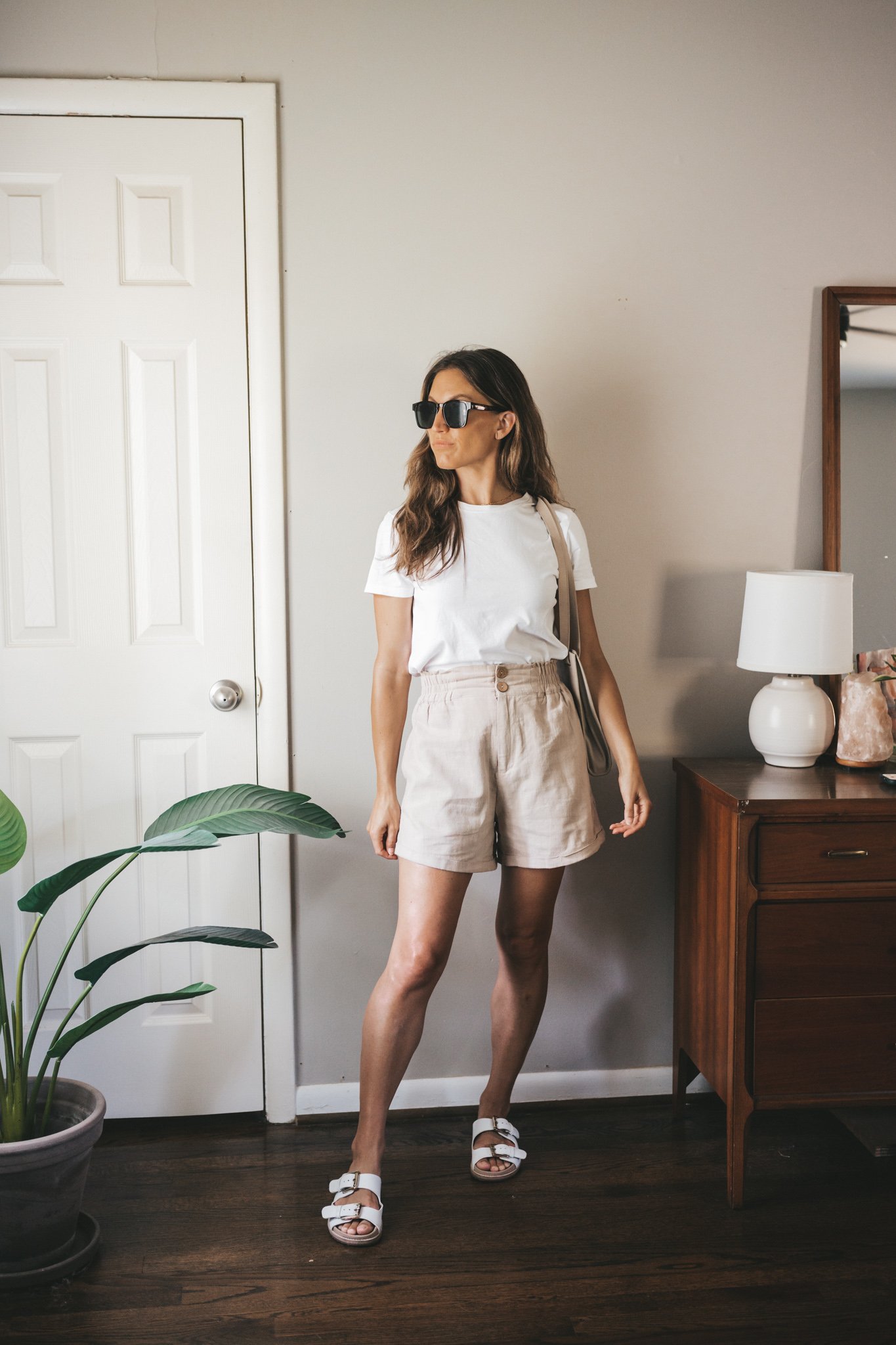 ashley wearing a white tee, tan linen shorts, white sandals, a gray shoulder bag, and tortoise shell sunglasses