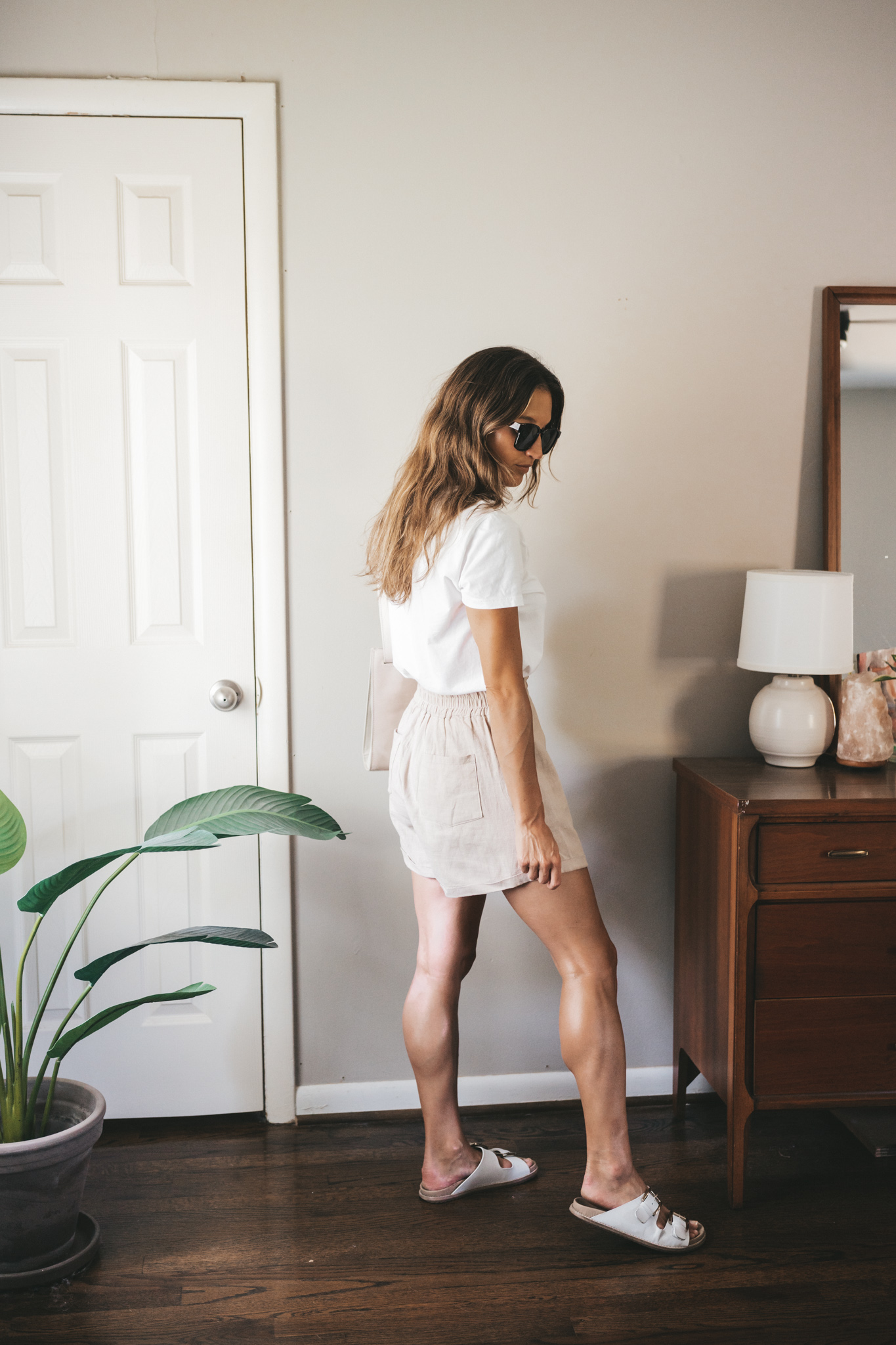 ashley wearing a white tee, tan linen shorts, white sandals, a gray shoulder bag, and tortoise shell sunglasses