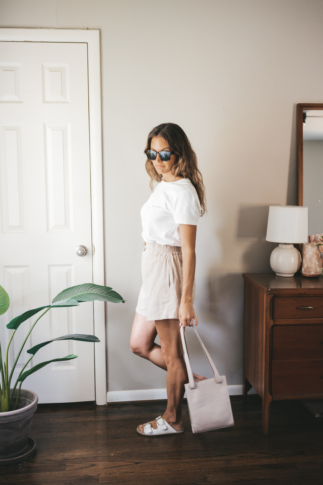 ashley wearing a white tee, tan linen shorts, white sandals, a gray shoulder bag, and tortoise shell sunglasses