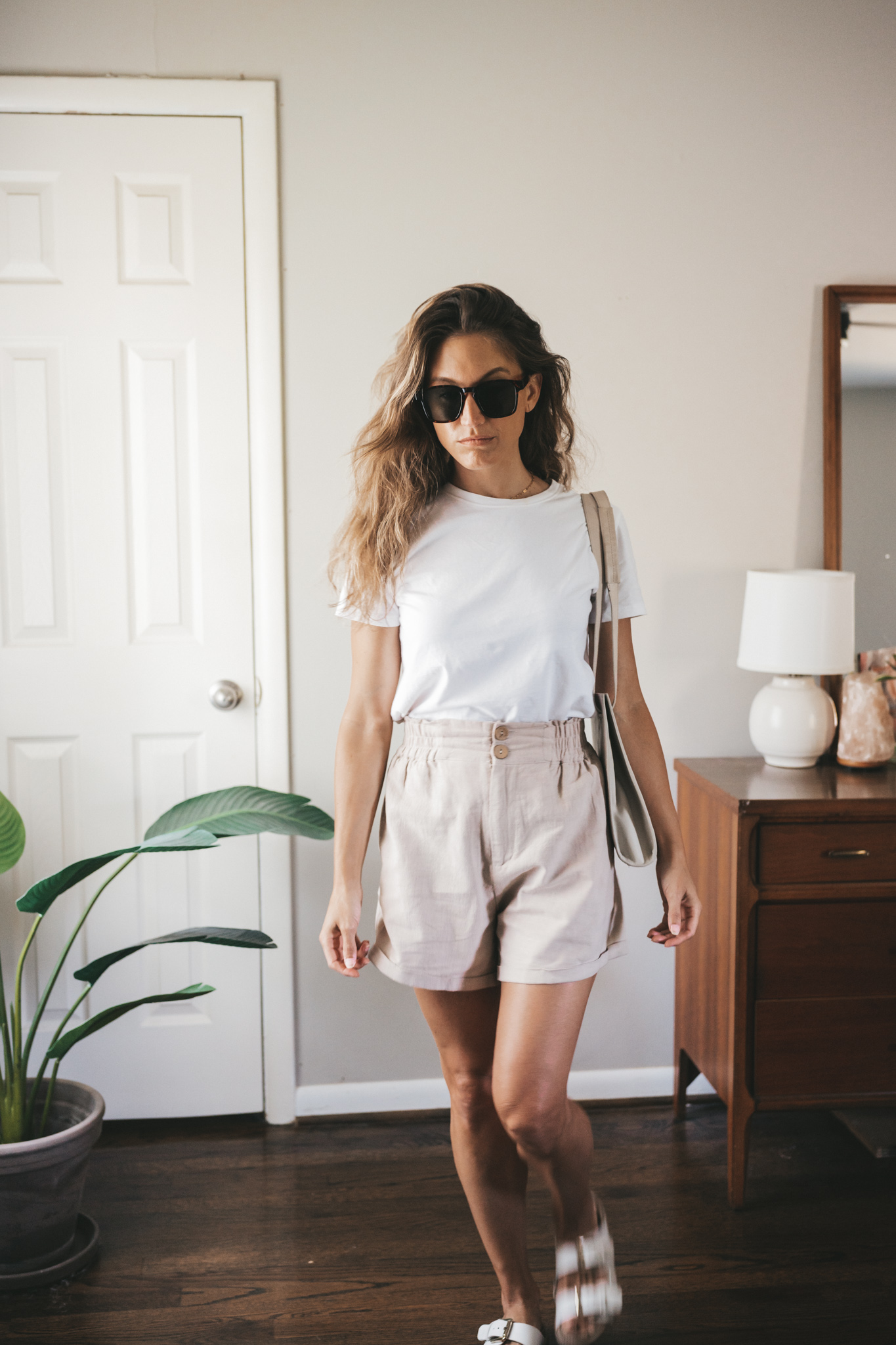 ashley wearing a white tee, tan linen shorts, white sandals, a gray shoulder bag, and tortoise shell sunglasses