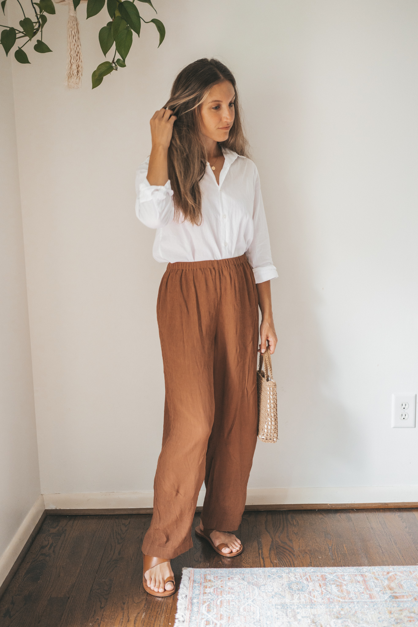 ashley wearing a white button down, brown drapey pants, brown leather sandals, and a woven bag