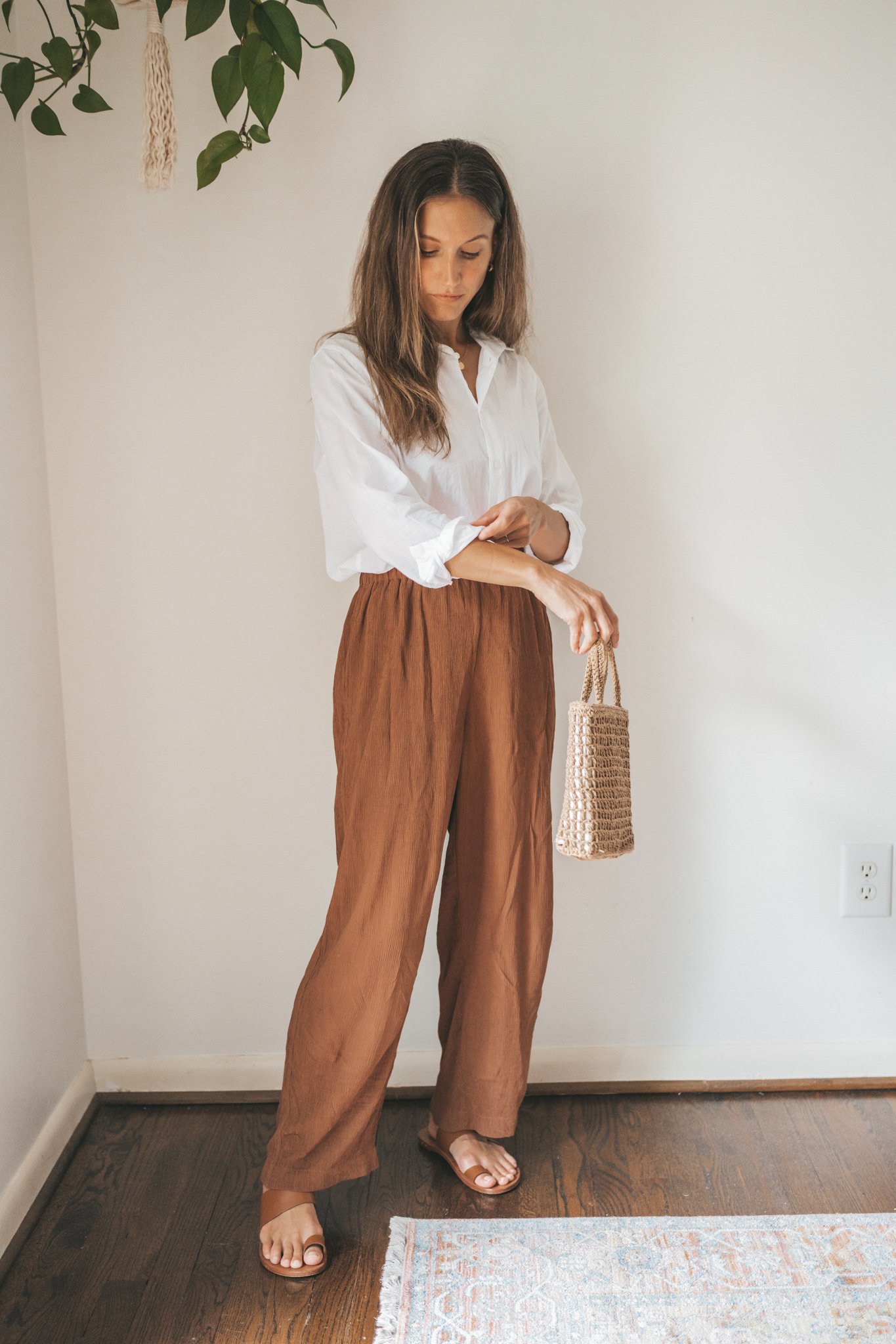ashley wearing a white button down, brown drapey pants, brown leather sandals, and a woven bag